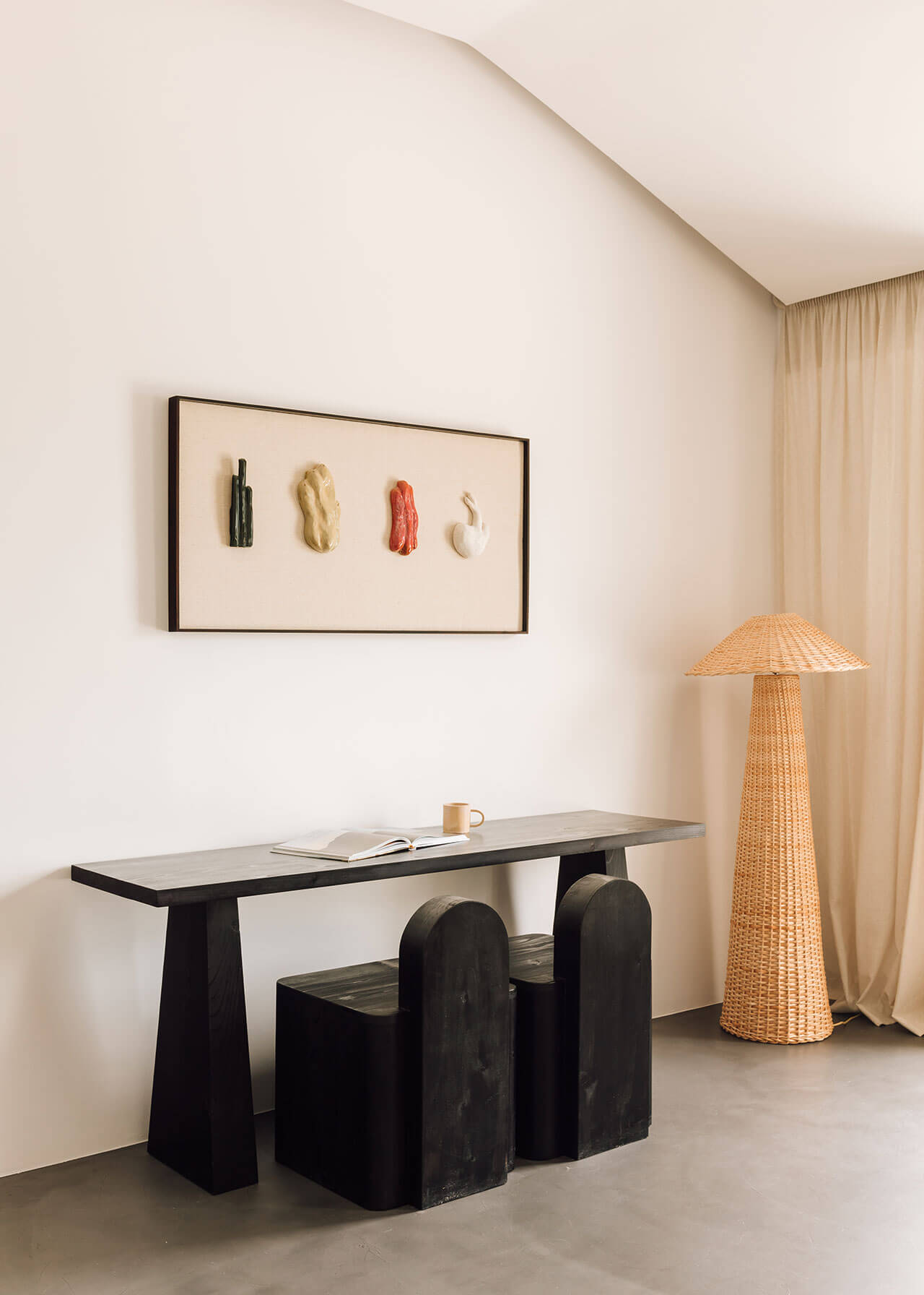 White Hotel, Europe — Minimalist workspace with black sculptural desk, woven rattan floor lamp, framed botanical artwork, and cream curtains.
