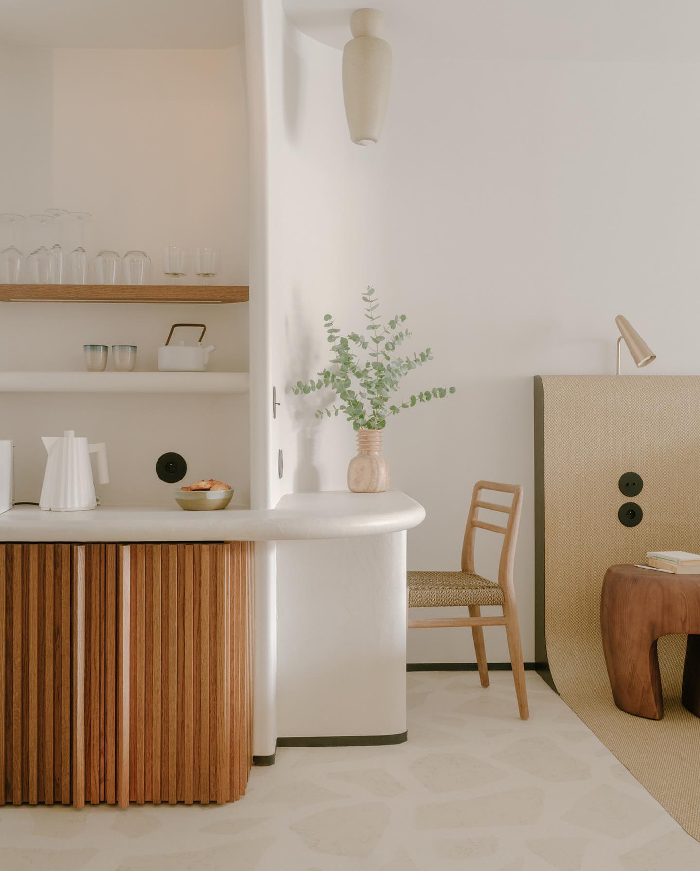 Belle Plage, Europe — Minimalist hospitality interior with curved white counter, vertical wood paneling, woven chair, potted eucalyptus, and soft neutral palette.