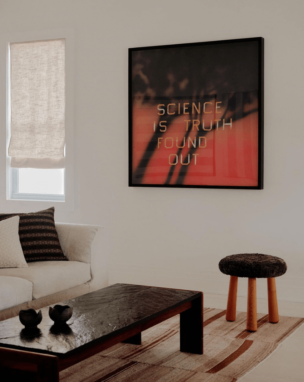 The James Bradley, North America — Modern living room with cream sofa, dark coffee table, mid-century stool, and framed neon artwork on pale wall beside linen window shade.