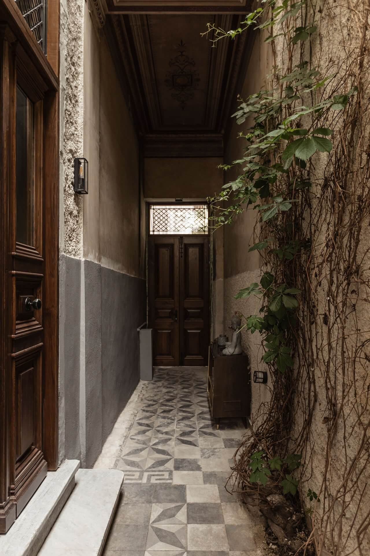 Casa Somos Suenos, Europe — Narrow ivy-covered passageway with geometric tile flooring, wooden doors, and natural light from transom window.