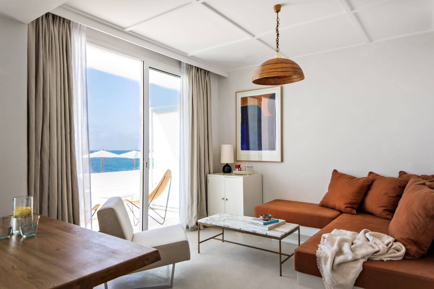 Les Roches Rouges, Europe — Coastal suite with terracotta sofa, wooden pendant lamp, marble coffee table, and sea-view balcony with white umbrella.