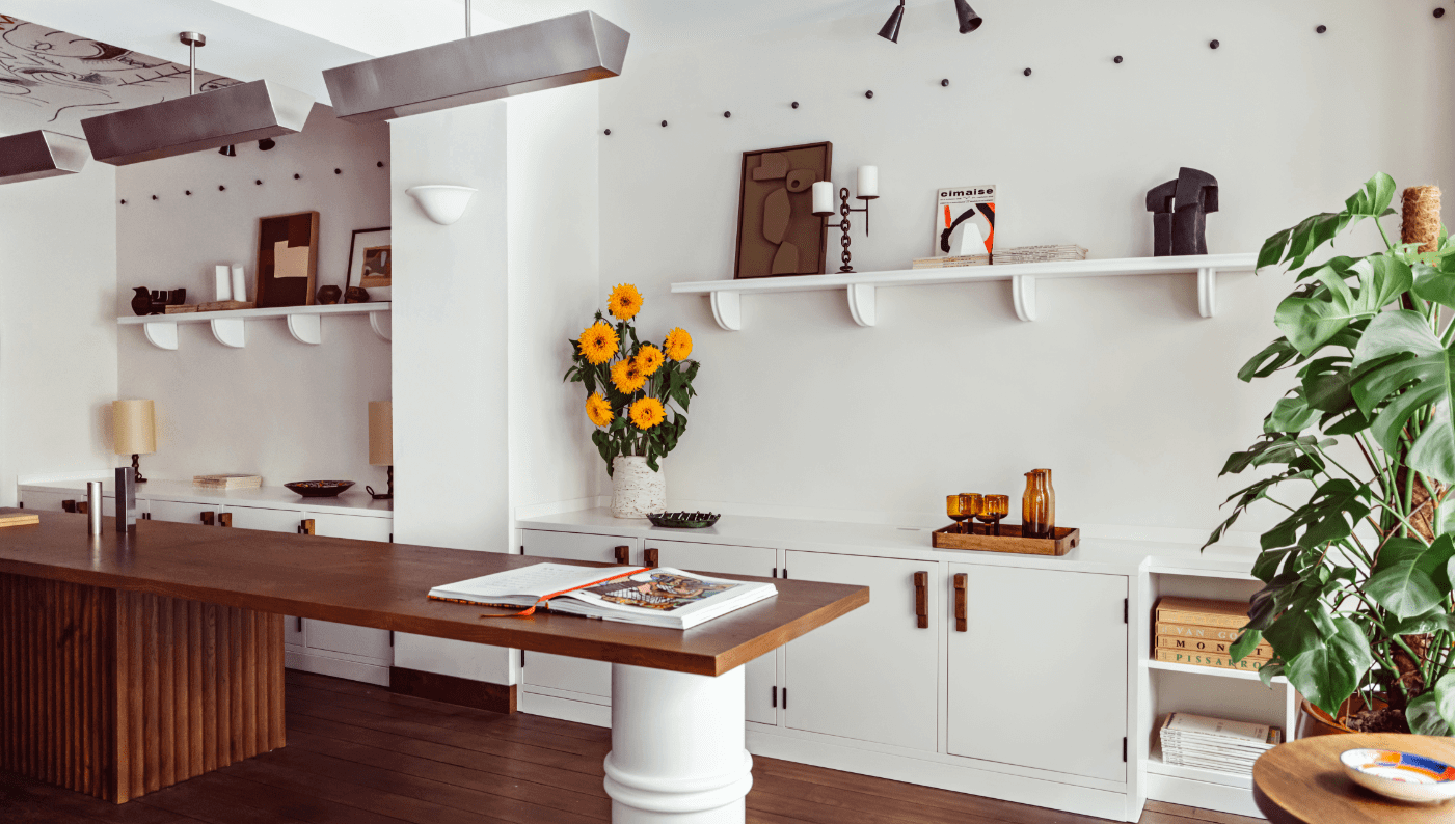 Académies et des Arts, Europe — Modern kitchen with walnut island, white cabinetry, floating shelves, brass accents, yellow gerberas, potted plants, and warm minimalist design.