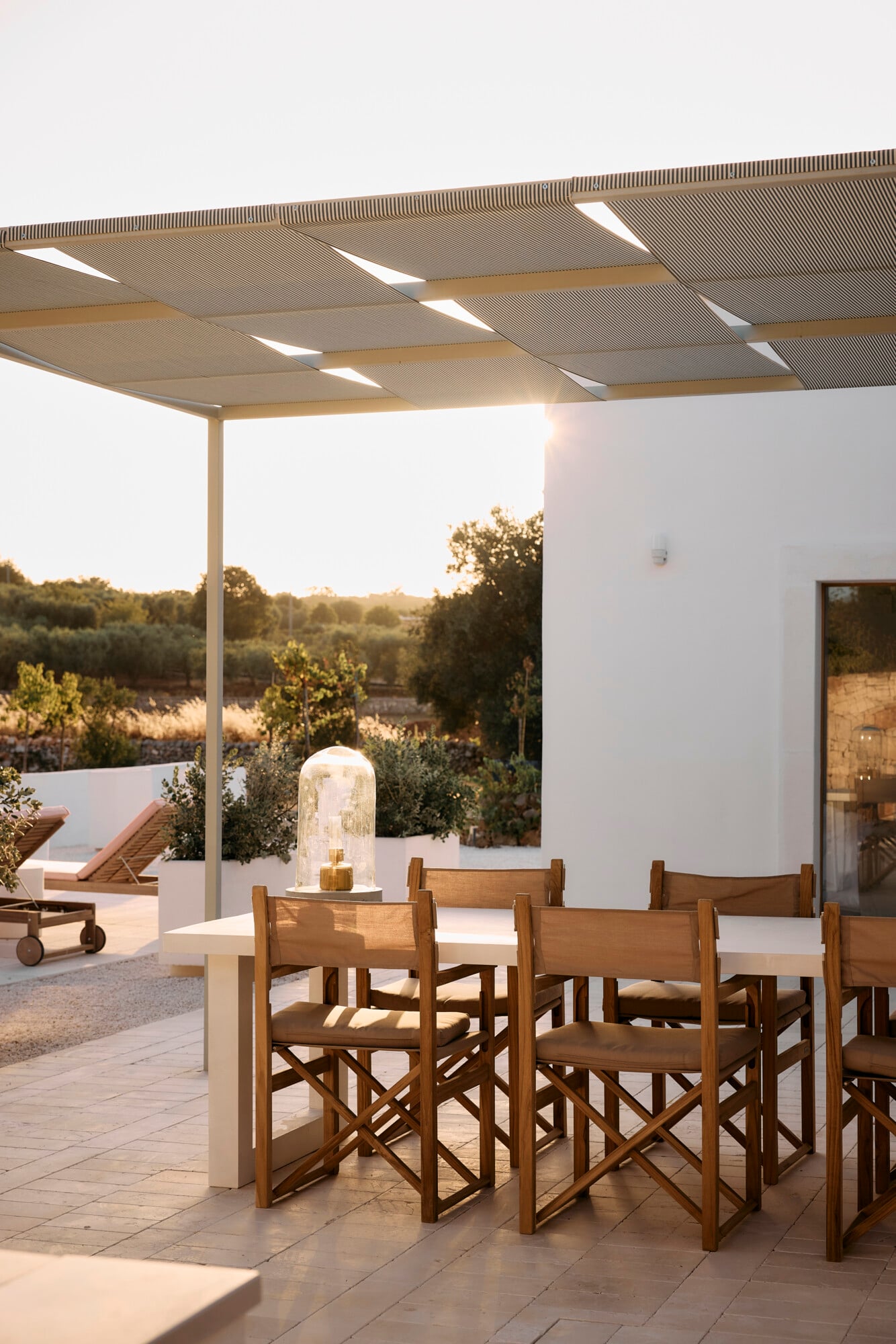 Villa Sessana, Italy — Modern pergola with integrated lighting shelters a dining table and wooden chairs overlooking olive groves at sunset.