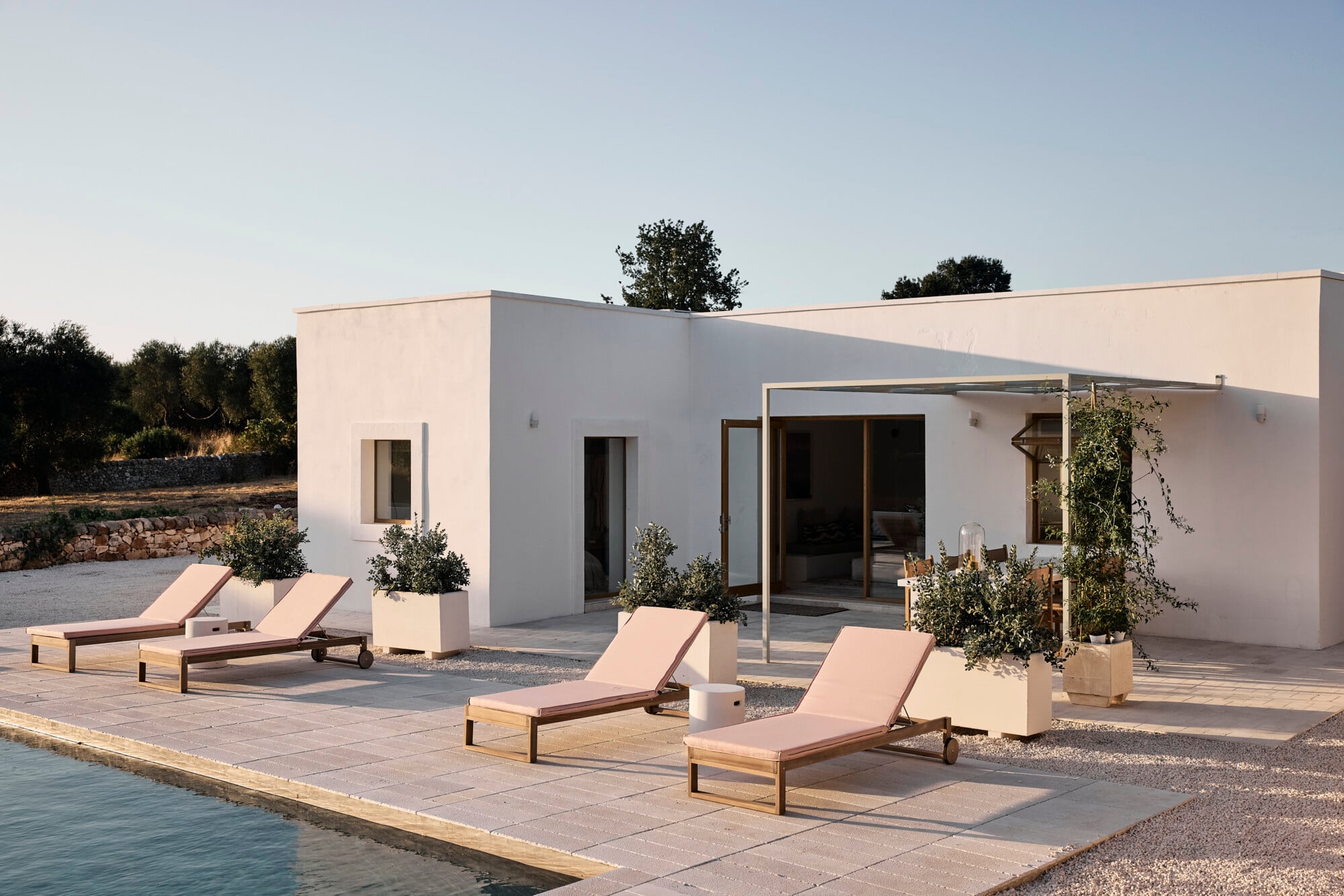 Villa Sessana, Italy — Modernist waterfront villa with crisp white architecture, timber loungers on pale stone terrace, potted olive trees, and natural Mediterranean light at dusk.