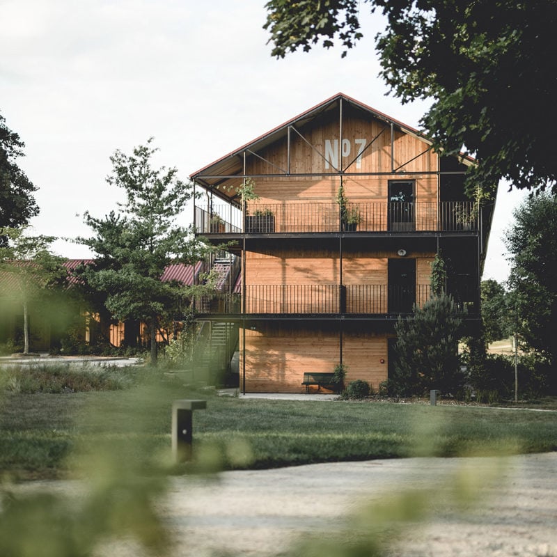 Le Barn, Europe — Three-story timber barn with black metal railings and large windows, surrounded by manicured lawn and mature trees, architectural photography at golden hour.