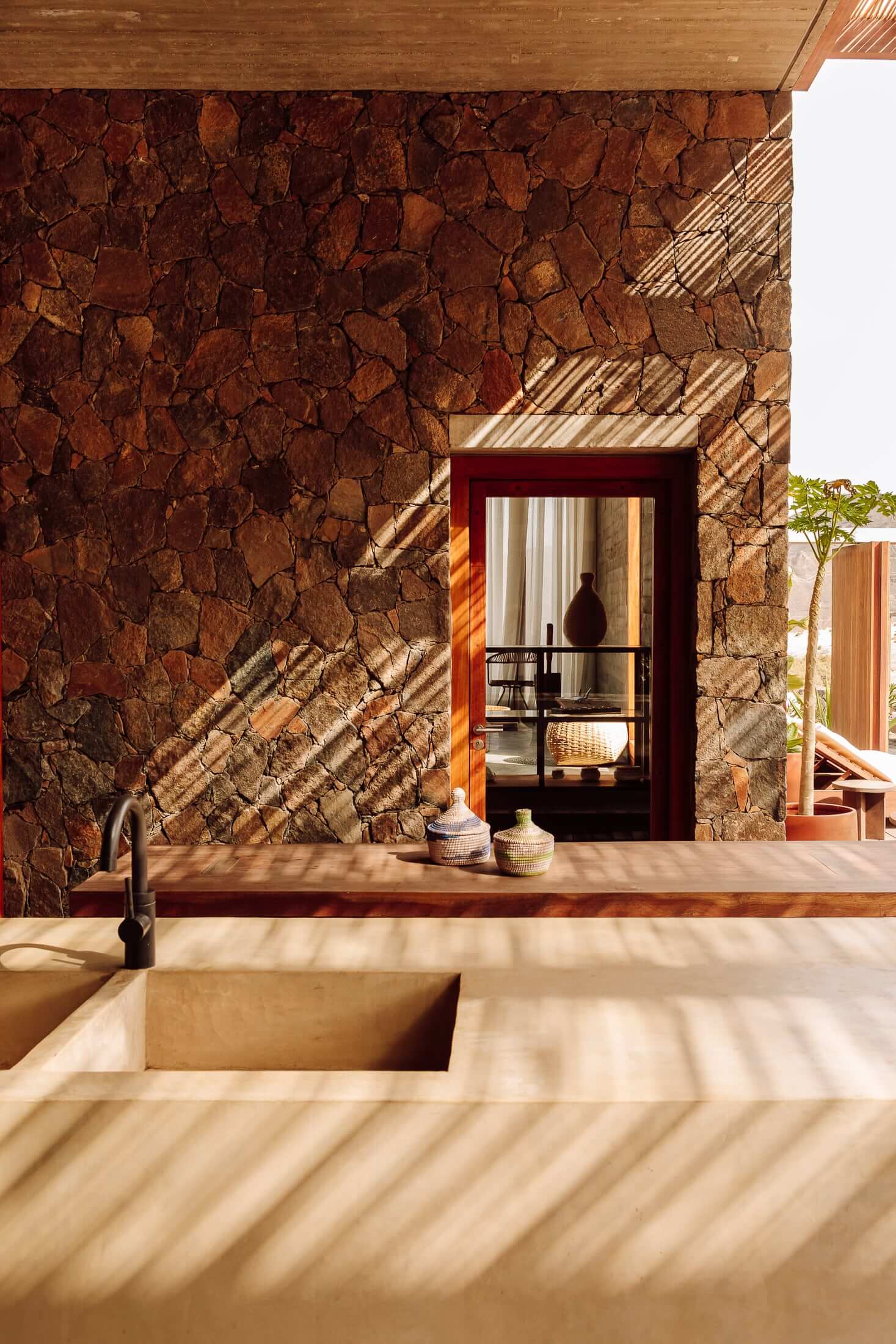 Barefoot Luxury, Africa — Warm stone wall, minimalist sink counter, and open doorway with afternoon light casting geometric shadows across terracotta surfaces.