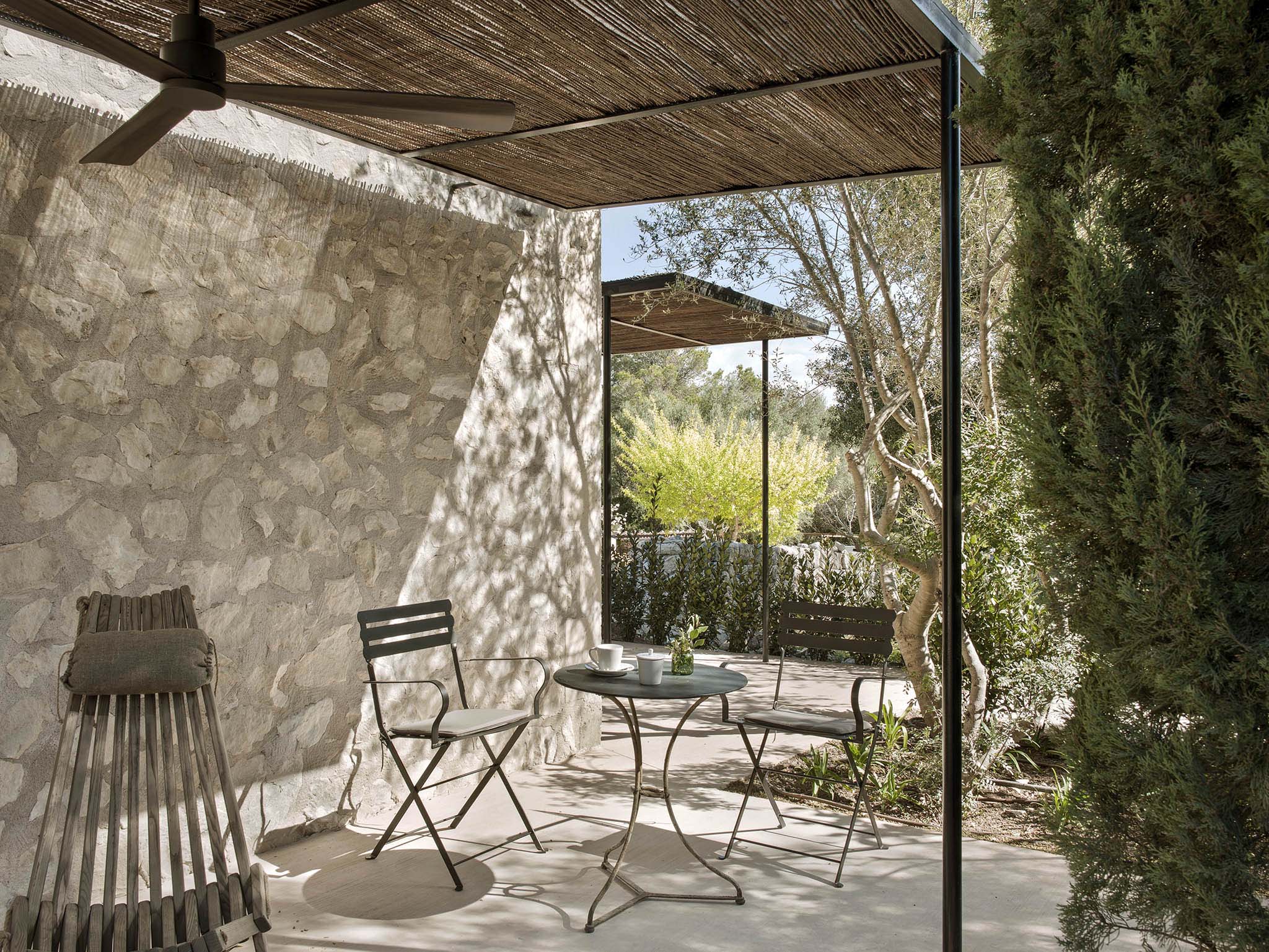 Finca Serena, Balearic Islands, Spain — Contemporary terrace with woven pergola, stone walls, metal chairs, and Mediterranean garden views casting dappled shadows in afternoon light.