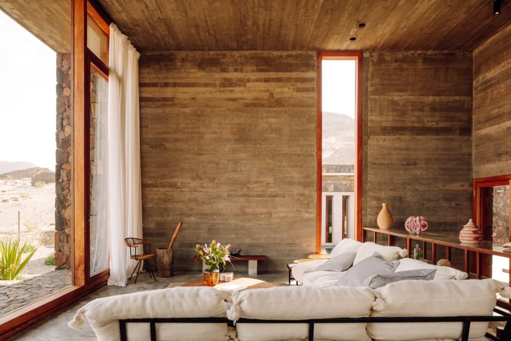 Barefoot Luxury, Africa — Minimalist bedroom with rammed earth walls, wooden ceiling, metal bed frame, white linens, and open doors to desert landscape.
