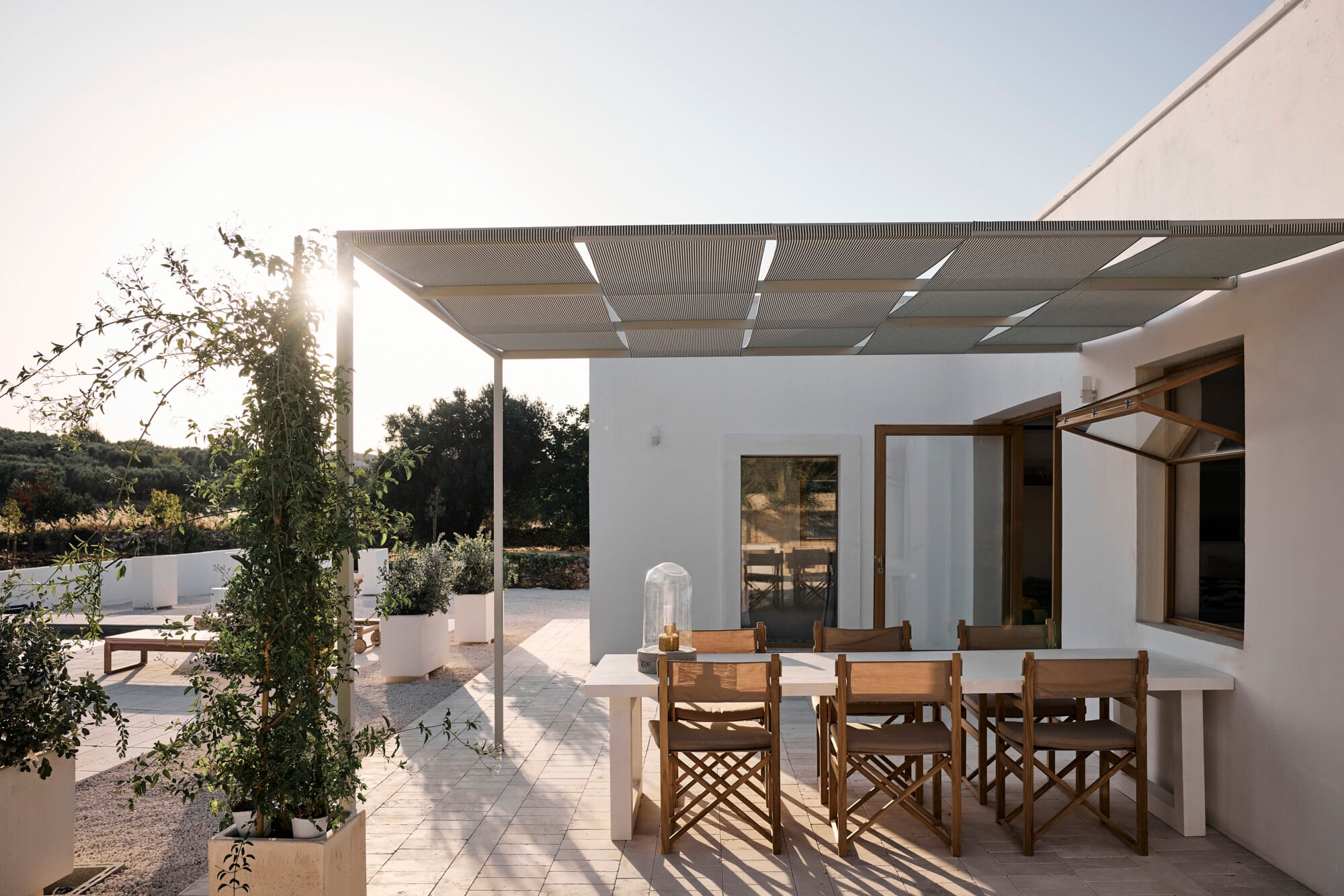 Villa Sessana, Italy — Modern white terrace with wooden pergola, dining table, natural light, potted plants, and Mediterranean landscape views.