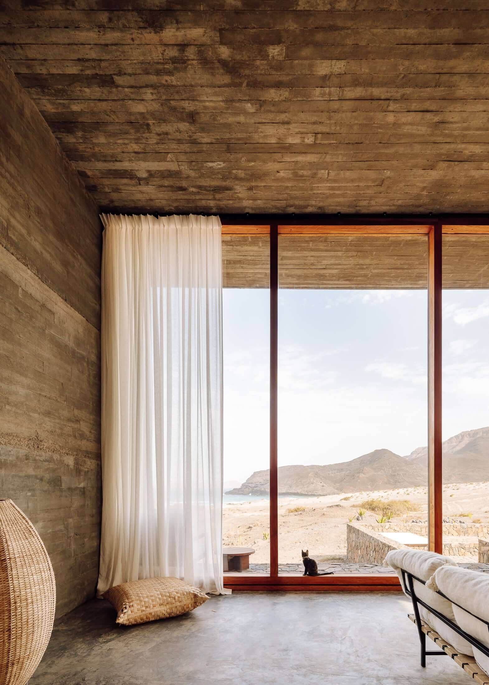 Barefoot Luxury, Africa — Concrete bedroom with wooden ceiling, sheer curtains, and expansive desert landscape view through floor-to-ceiling windows.