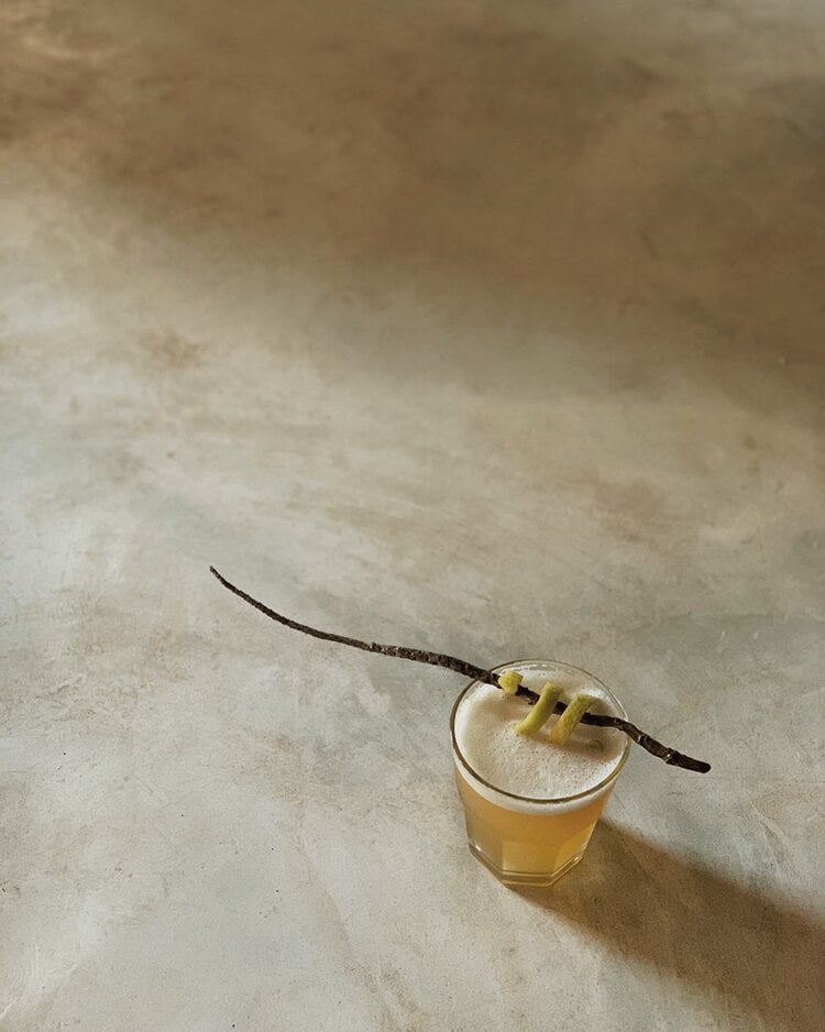 Sussurro, Africa — Craft cocktail with vanilla bean garnish and two-tone layered drink on concrete surface, natural light casting shadow.