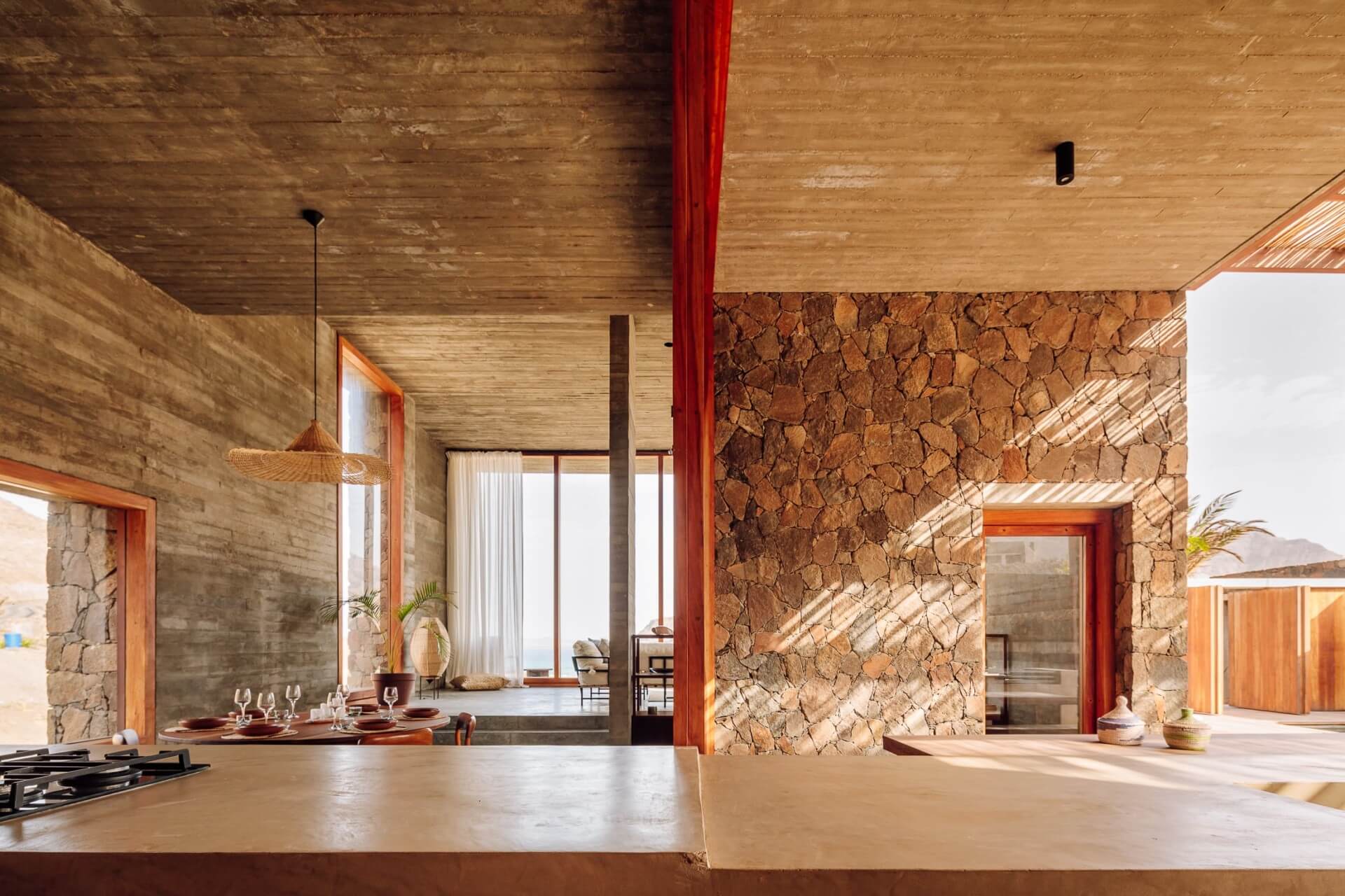Barefoot Luxury, Africa — Open-plan interior with cork walls, wooden ceiling, red steel columns, concrete floors, and natural light casting geometric shadows across dining and living spaces.
