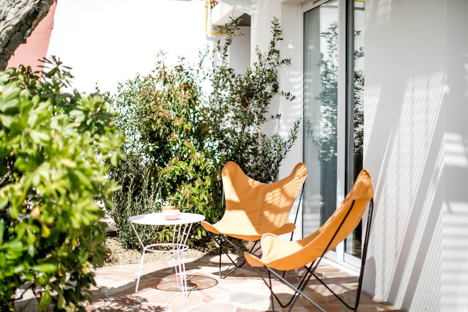 Les Roches Rouges, Europe — Terracotta butterfly chairs in dappled sunlight on minimalist courtyard with potted greenery and modernist windows.