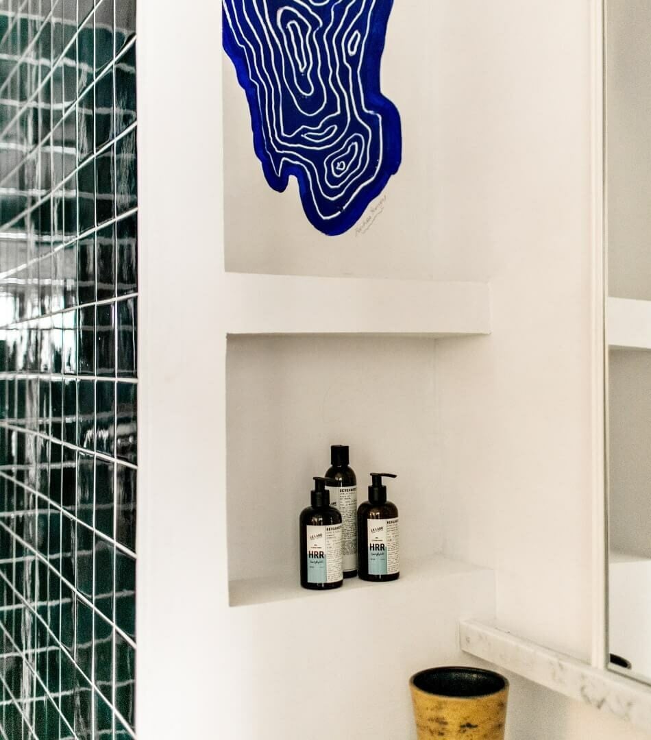 Les Roches Rouges, Europe — Modern bathroom niche with blue topographic wall art, dark glass tile, black skincare bottles, and ochre ceramic vessel.