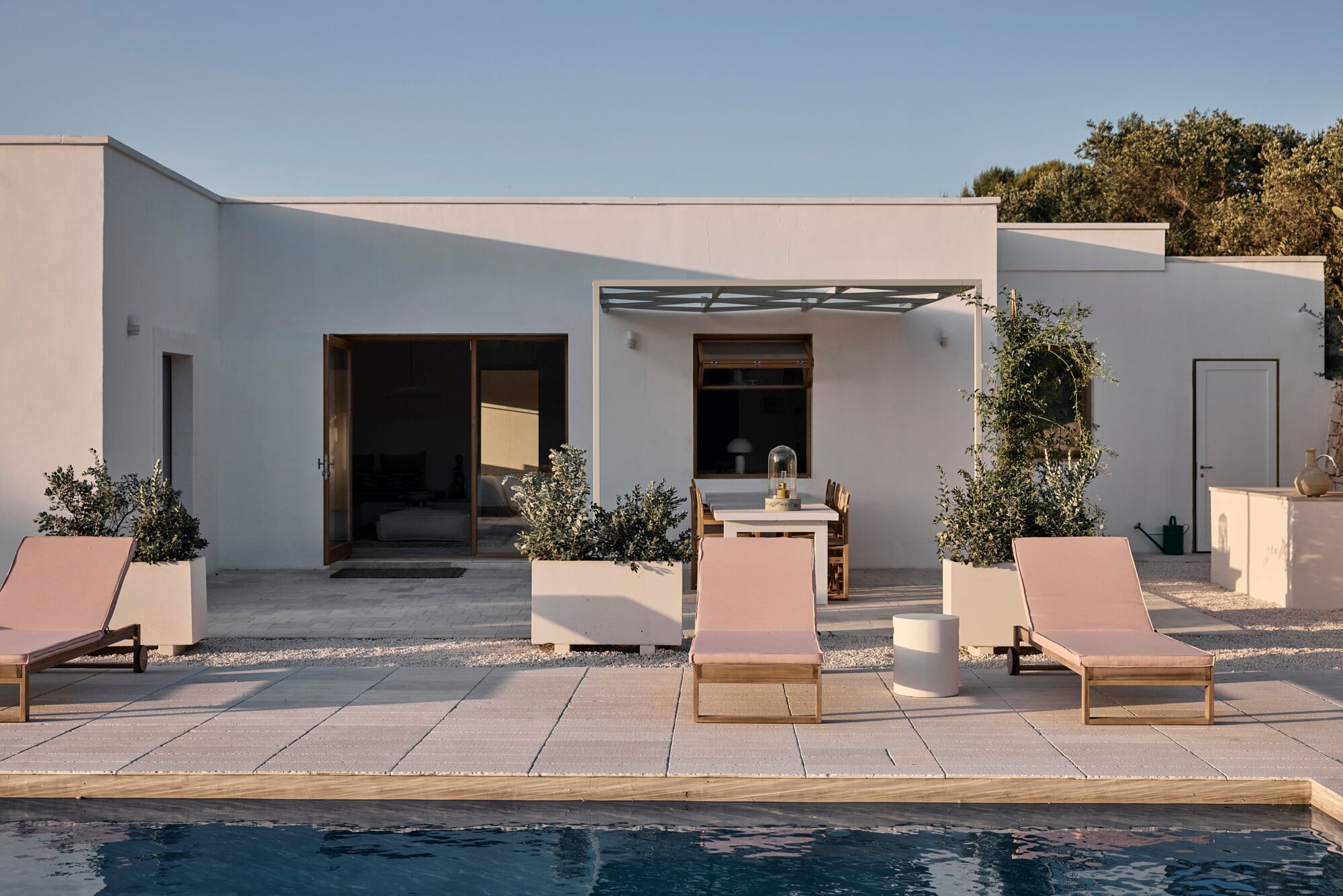 Villa Sessana, Italy — Minimalist poolside terrace with pink loungers, white rendered architecture, potted olive trees, and stepped roofline at golden hour.