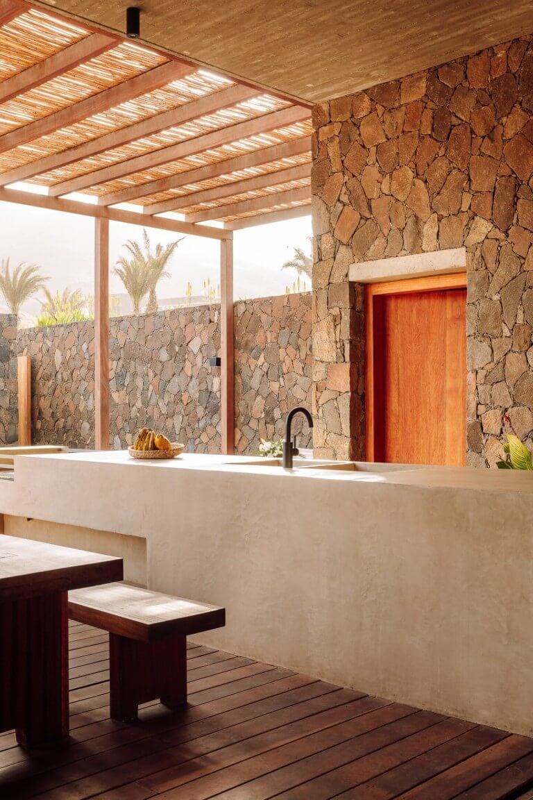 Barefoot Luxury, Africa — Rooftop kitchen with stone walls, wooden pergola, concrete counter, dark timber stools, and desert views at sunset.