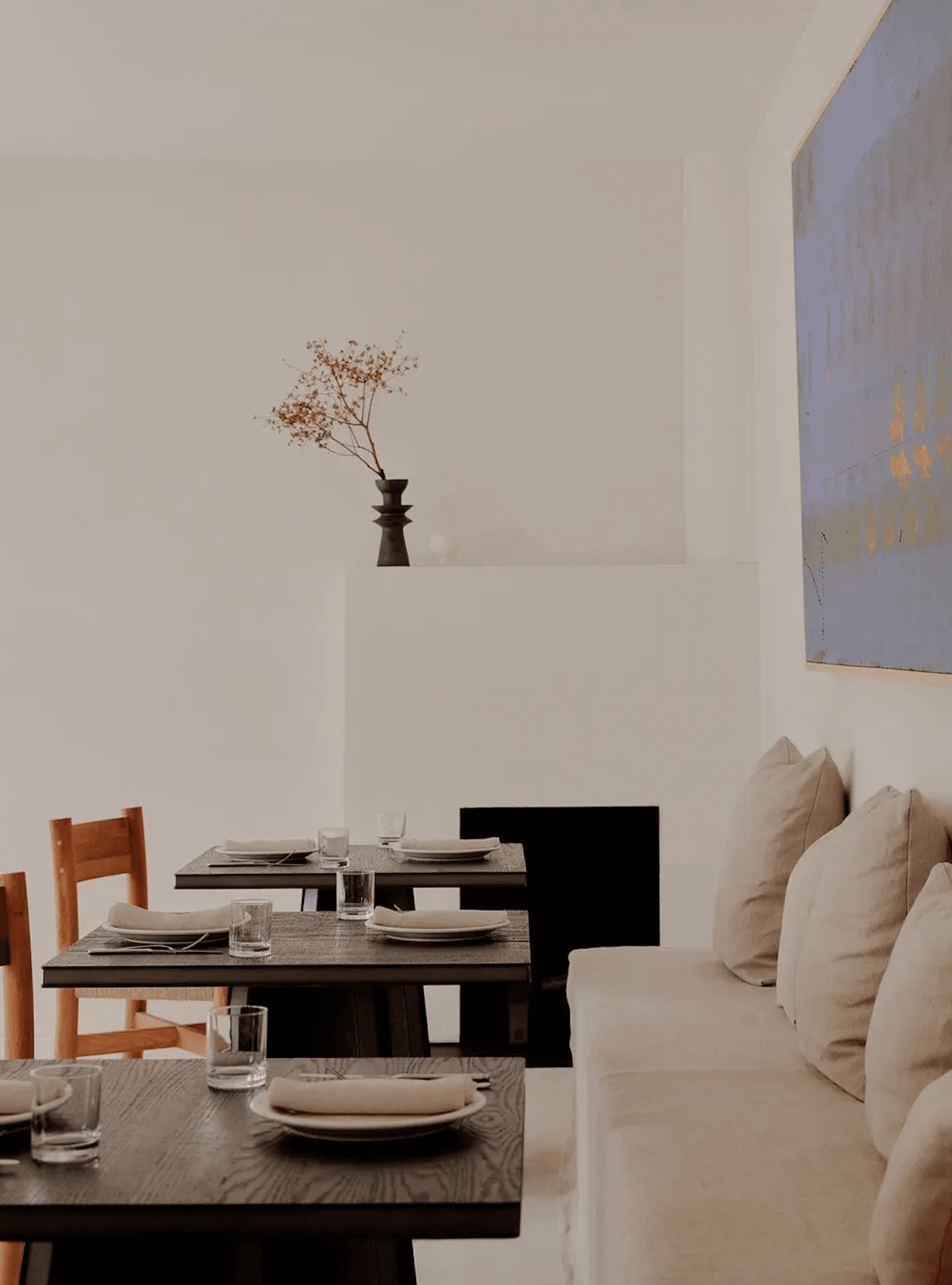 The James Bradley, North America — Minimalist dining room with black tables, cream upholstered seating, wooden chairs, dried botanicals in dark vase, and gallery artwork.