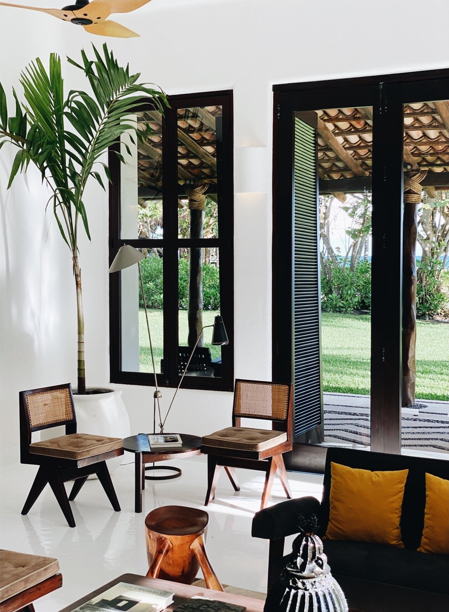 Hotel Esencia, Mexico — Modernist living room with black-framed glass doors, woven cane chairs, wooden side table, potted palm, and golden ceiling fan overlooking manicured garden.