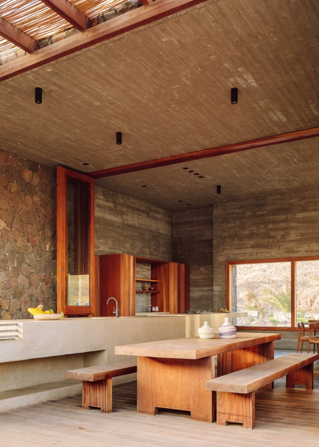 Barefoot Luxury, Africa — Open-plan dining and kitchen with wooden furniture, concrete ceiling, stone walls, and expansive views of natural landscape.