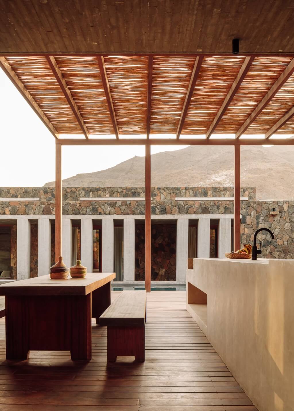 Barefoot Luxury, Africa — Woven timber pergola shelters minimalist kitchen pavilion with stone walls, wooden decking, and desert mountain views.