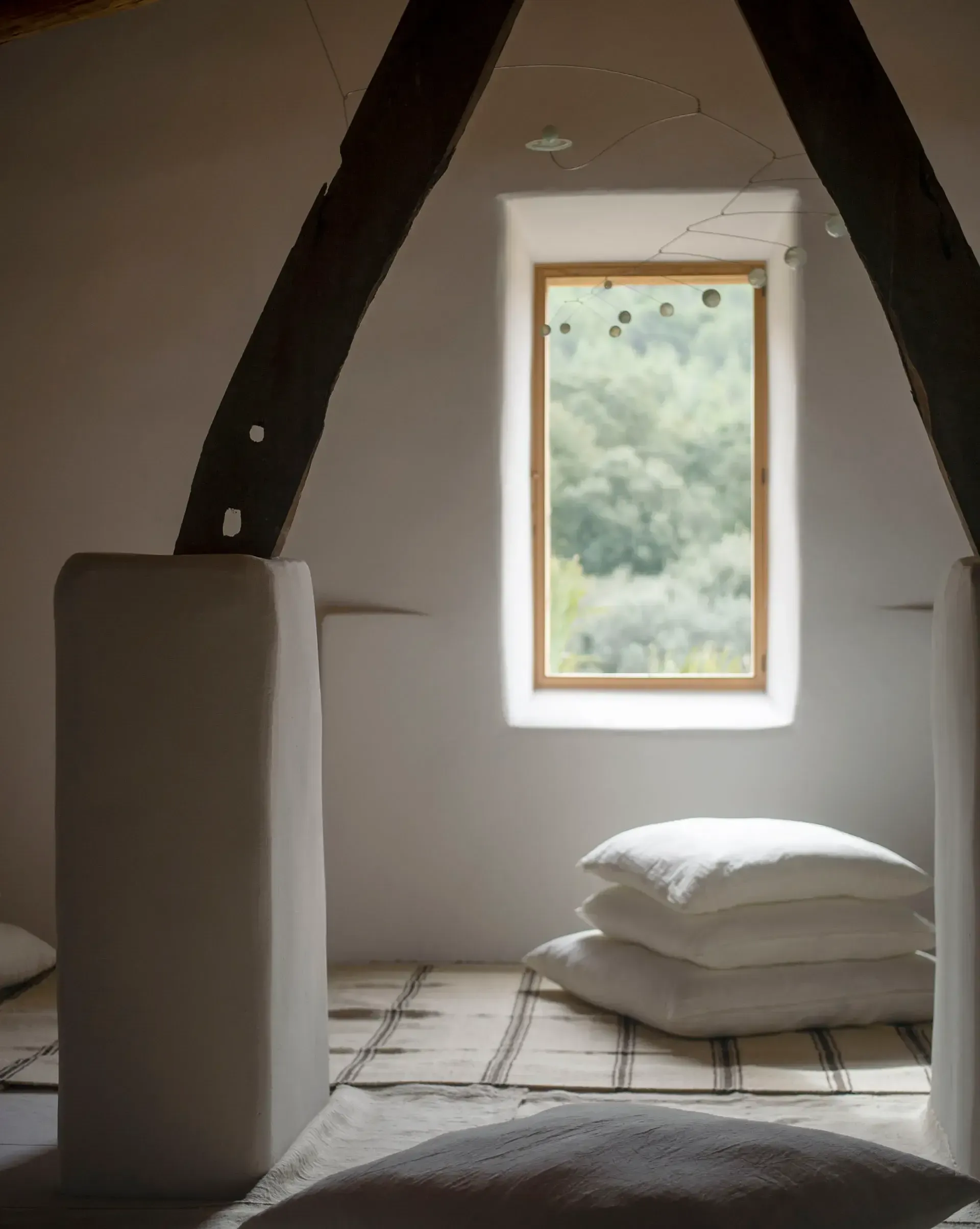 Es Racó d'Artà, Balearic Islands, Spain — Minimalist attic bedroom with dark timber beams, white walls, stacked pillows, and framed window overlooking forested landscape.