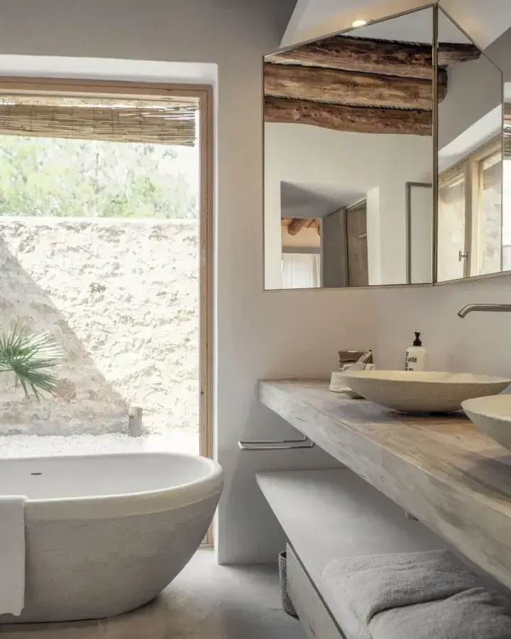 Es Racó d'Artà, Balearic Islands, Spain — Minimalist bathroom with reclaimed wood canopy mirror, stone vessel sinks, soaking tub, and limestone terrace opening to Mediterranean landscape.