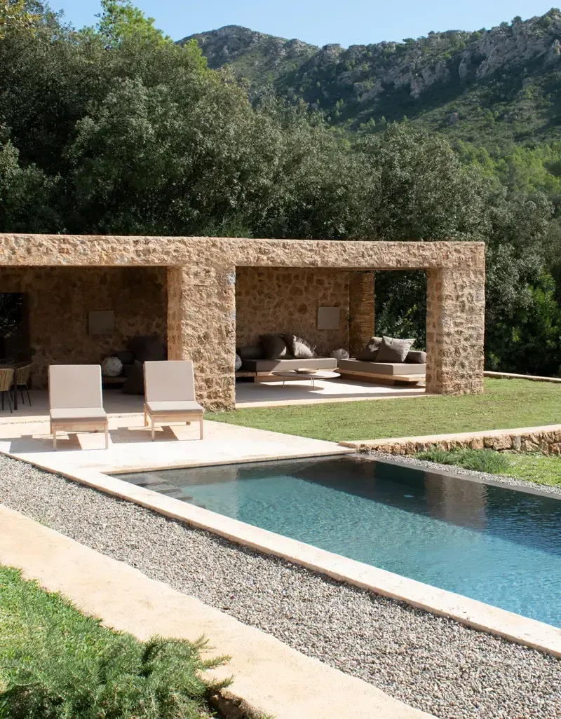 Es Racó d'Artà, Balearic Islands, Spain — Modernist stone pavilion with covered lounge overlooking rectangular pool, olive trees, and rocky mountainside in Mediterranean landscape.
