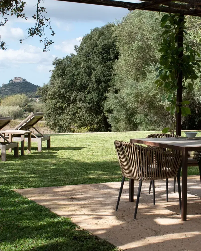 Es Racó d'Artà, Balearic Islands, Spain — Pergola-shaded terrace with rope chairs, olive trees, and hilltop castle views across manicured lawn.
