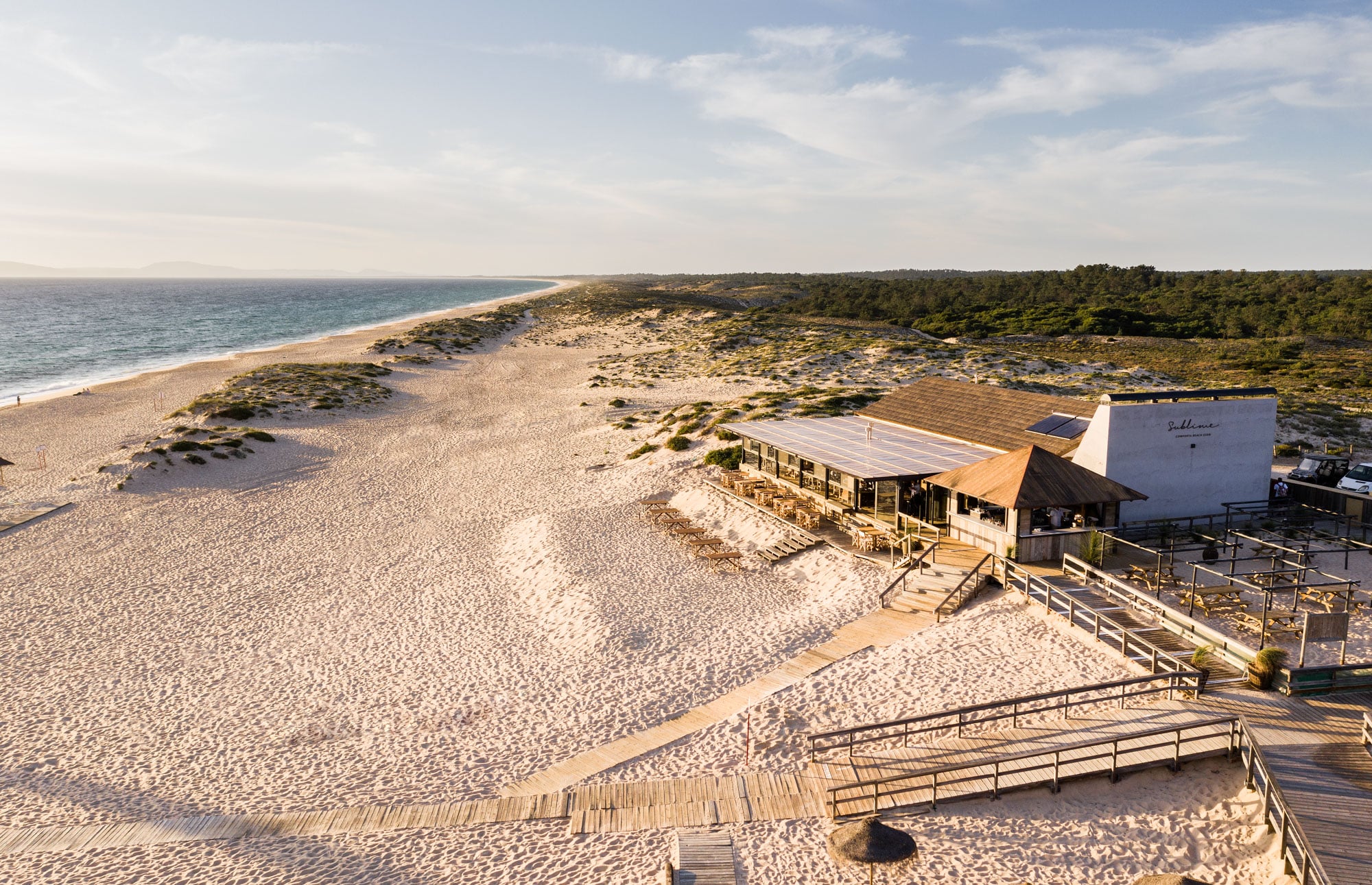 Sublime Comporta Beach Club, Portugal — Coastal pavilion with timber deck and metal railings overlooking sandy beach, dune vegetation, and turquoise sea under bright sky.