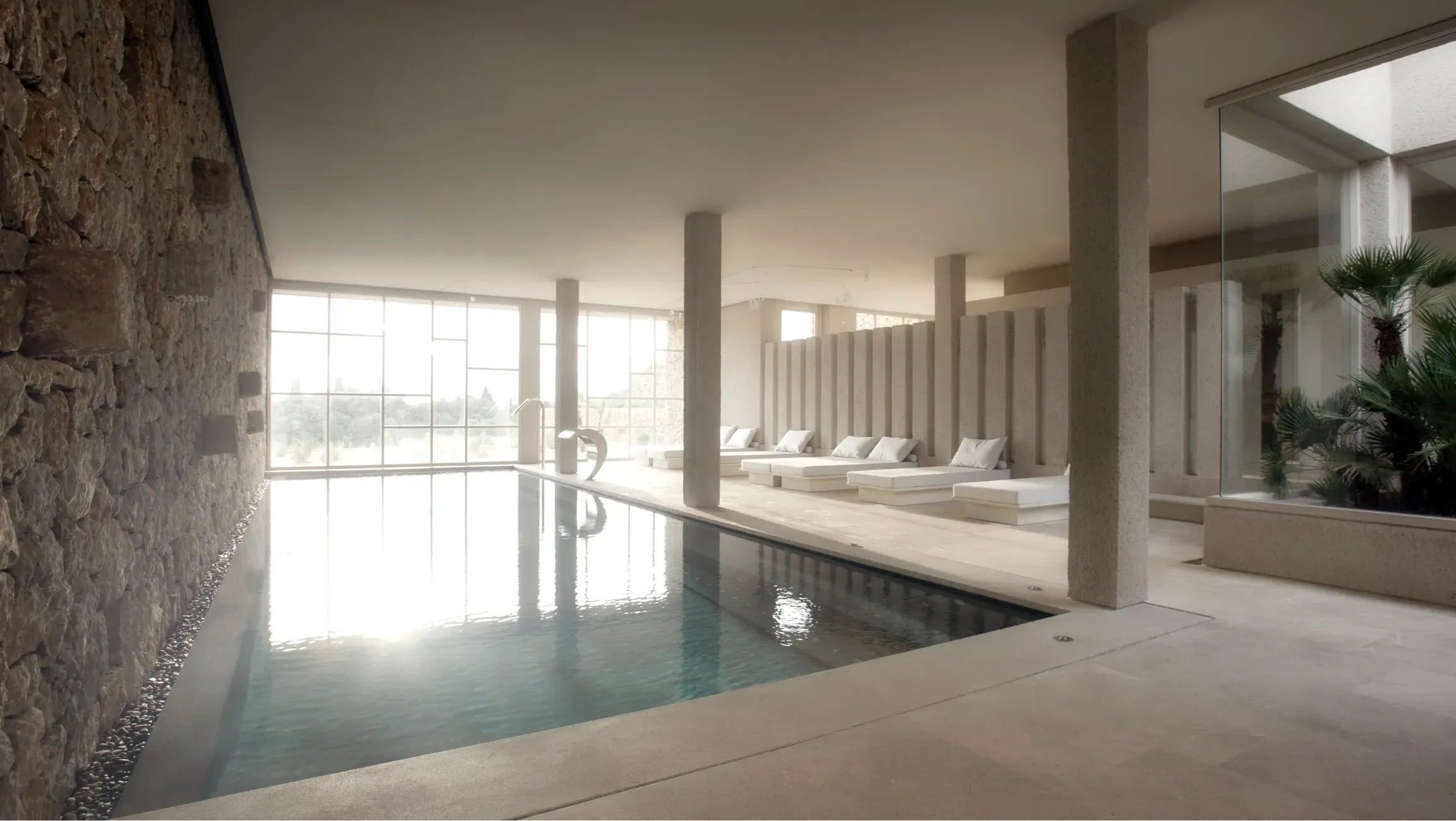 Es Raco Spa, Balearic Islands, Spain — Minimalist indoor pool with stone wall, concrete columns, and loungers reflecting architectural restraint and natural light.