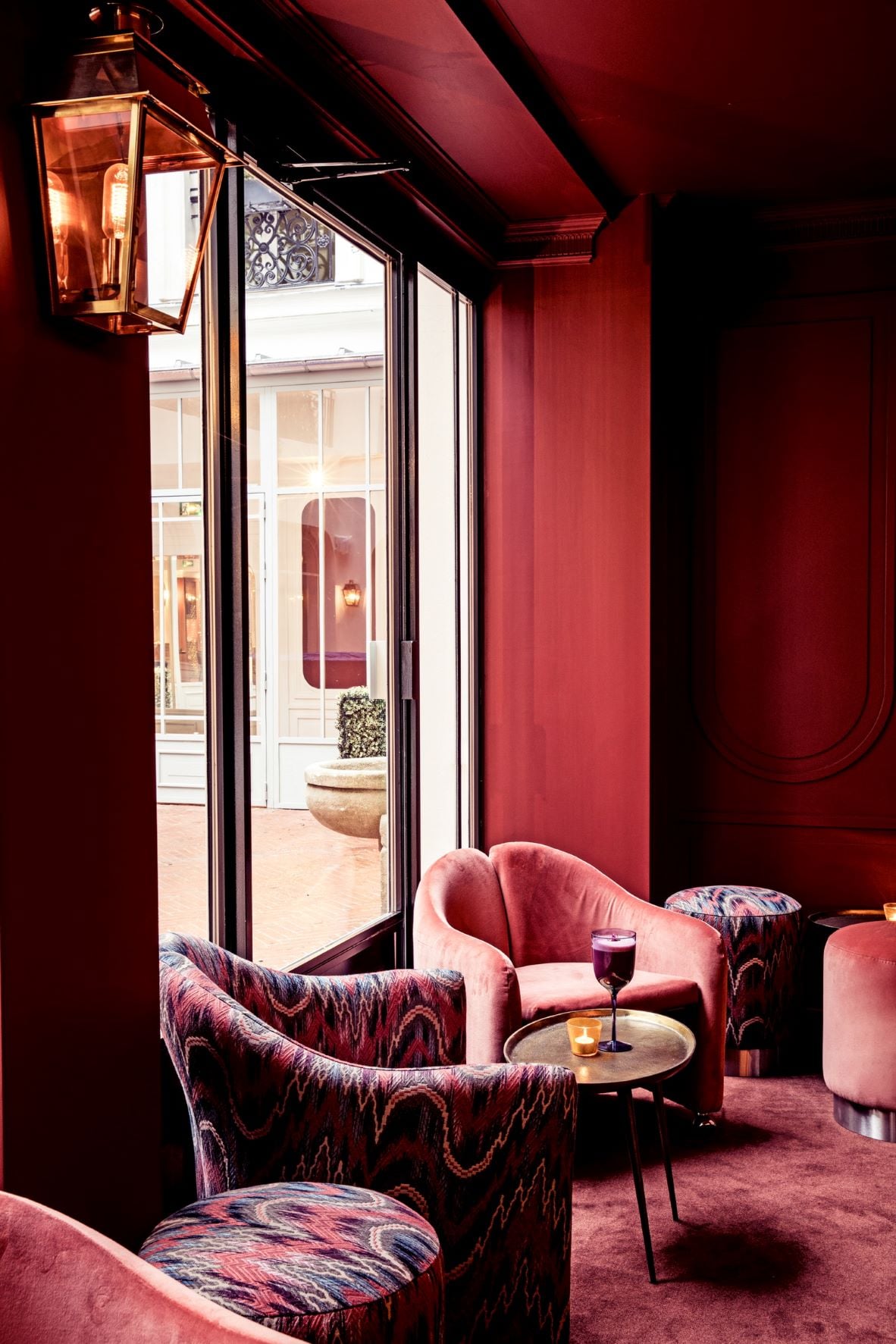 The Shell, Europe — Deep crimson lounge with geometric-patterned armchairs, brass side table, lantern sconce, and open French doors to sunlit courtyard.