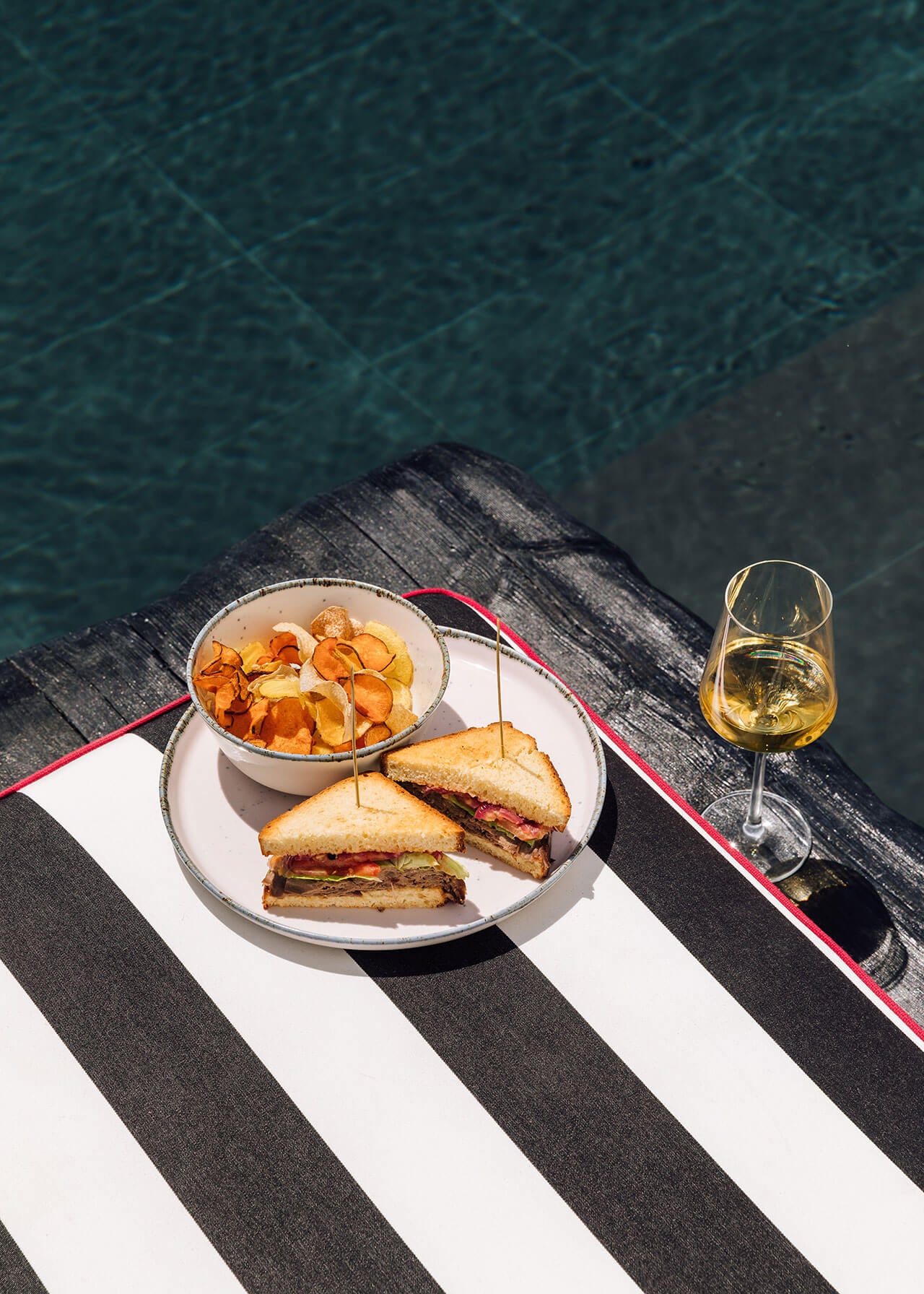 White Hotel, Europe — Poolside lunch setup with grilled sandwiches, crispy chips, and white wine overlooking turquoise water, striped lounger in foreground.