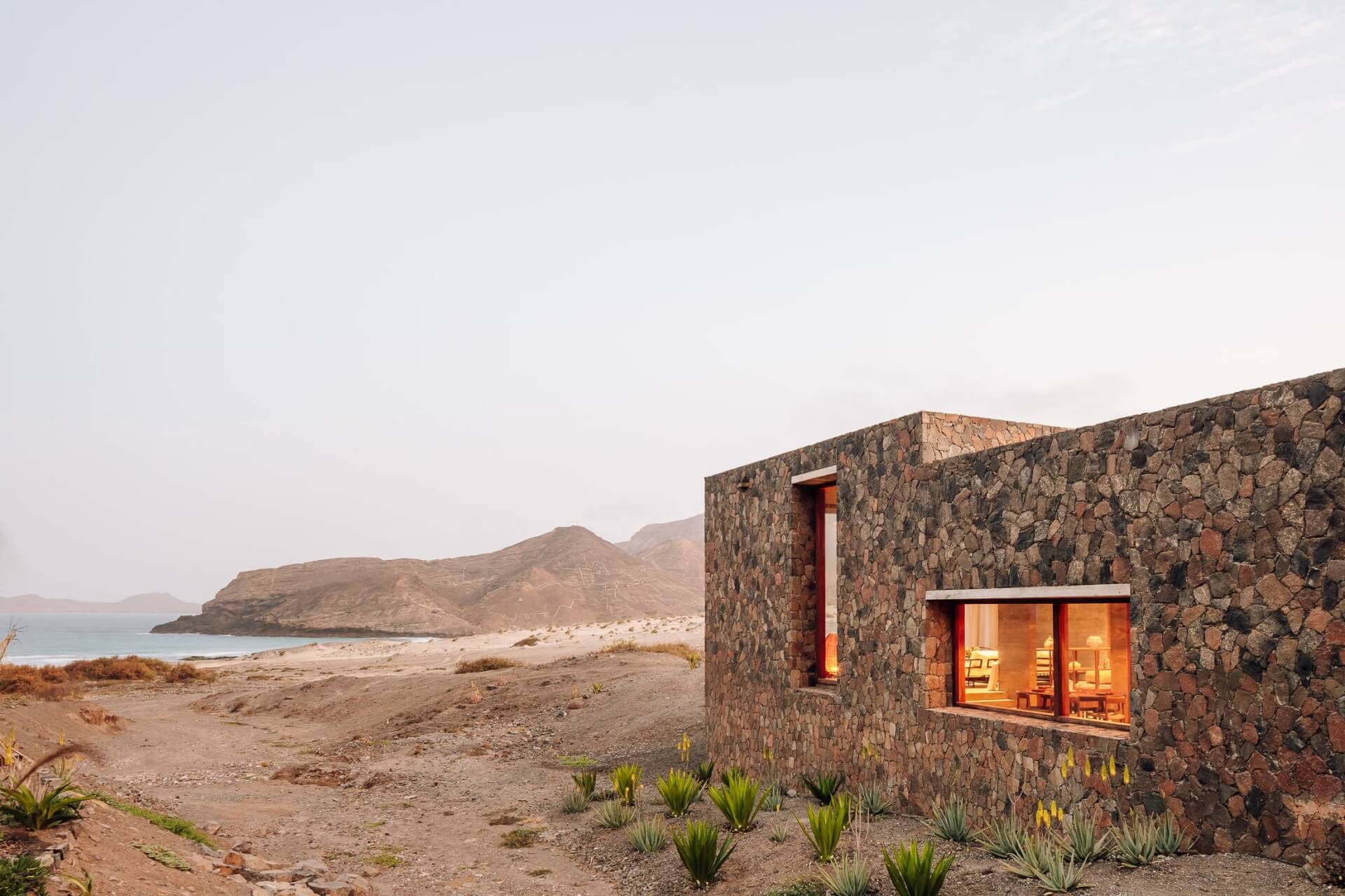 Barefoot Luxury, Africa — Stone pavilion with warm interior light overlooking desert beach and turquoise ocean at dusk, minimalist coastal architecture.