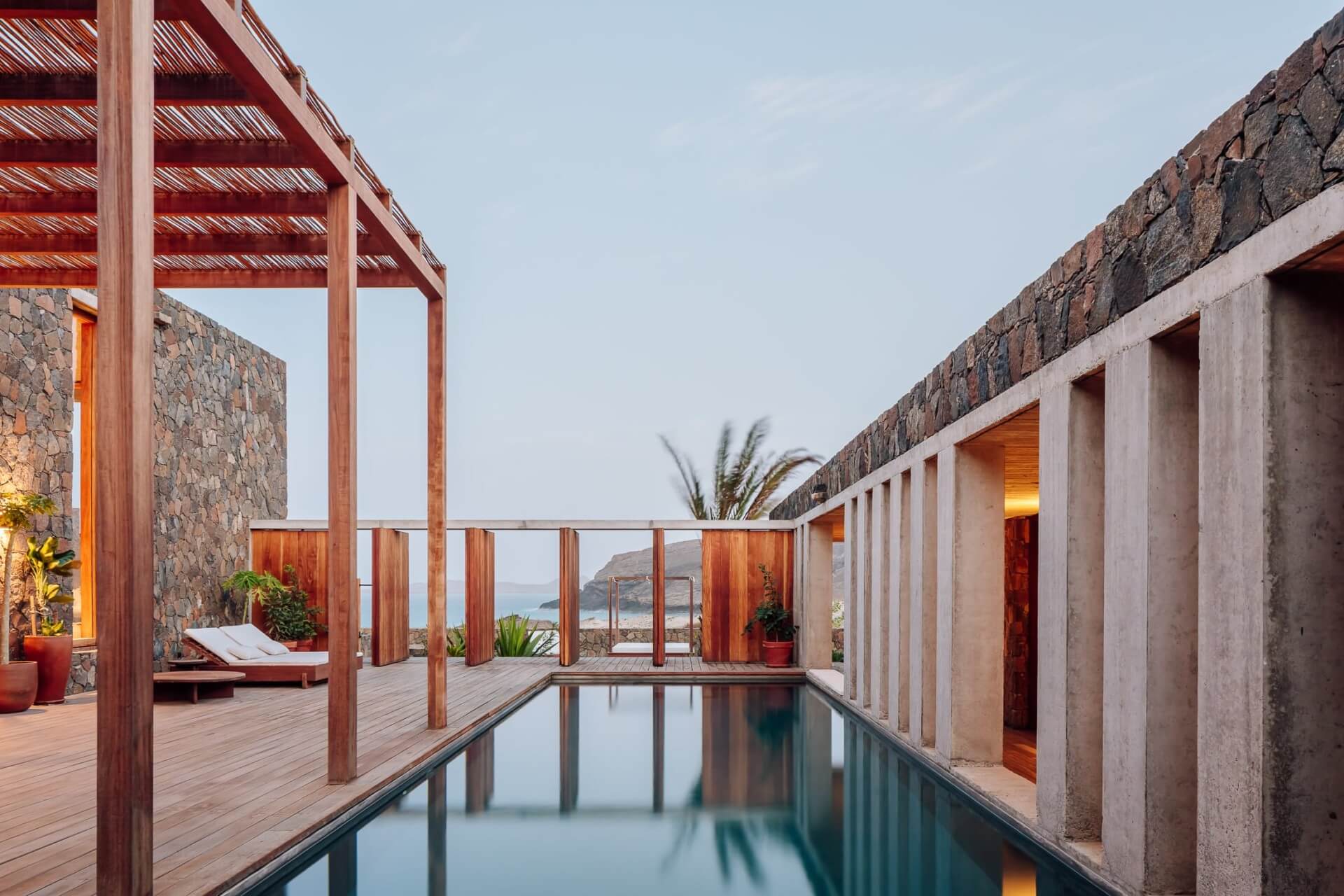 Barefoot Luxury, Africa — Minimalist poolside terrace with timber pergola, stone walls, and ocean views at dusk.