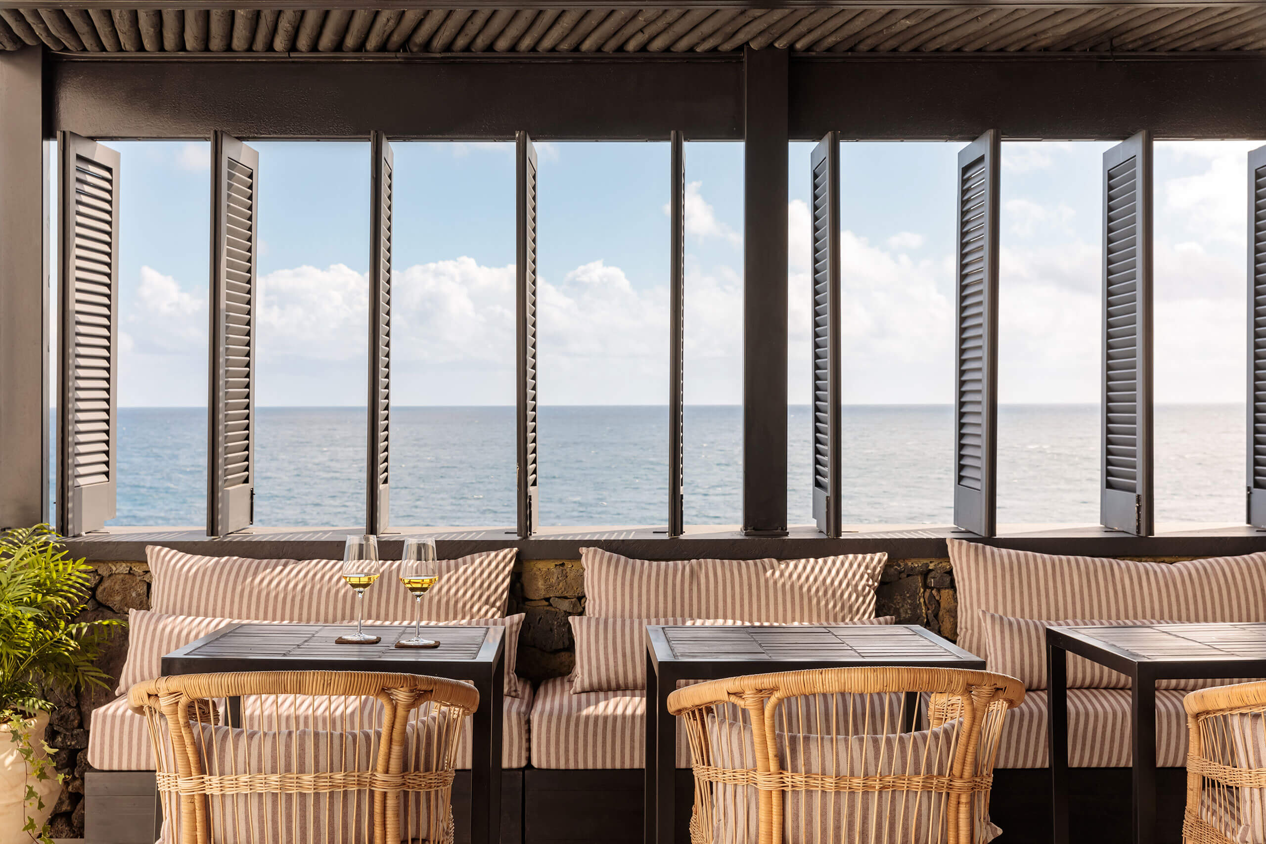 White Hotel, Europe — Coastal dining pavilion with louvered doors opening to sea view, woven chairs, striped cushions, and natural light.