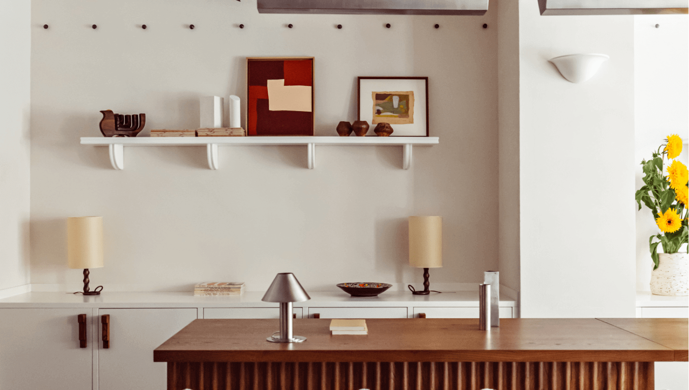 Académies et des Arts, Europe — Minimalist cream wall with floating shelf displaying red artwork, wooden desk with brass lamp, and warm natural light casting soft shadows.