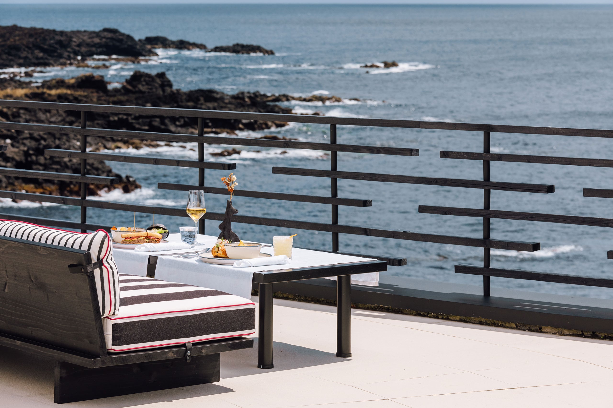 White Hotel, Europe — Coastal dining table set with white linens and wine glass overlooking rocky shoreline, modern black railing and striped lounge chair.