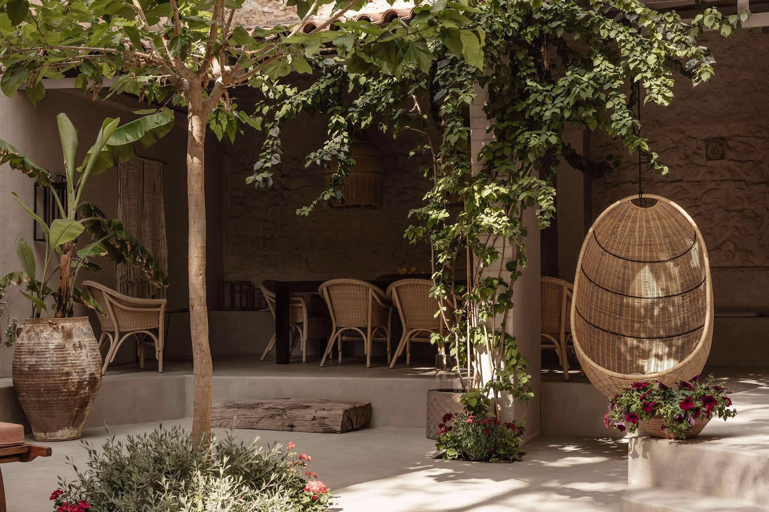 Casa Somos Suenos, Europe — Shaded courtyard with woven pendant chair, climbing vines, rattan planters, natural stone flooring, and scattered seating beneath dappled sunlight.