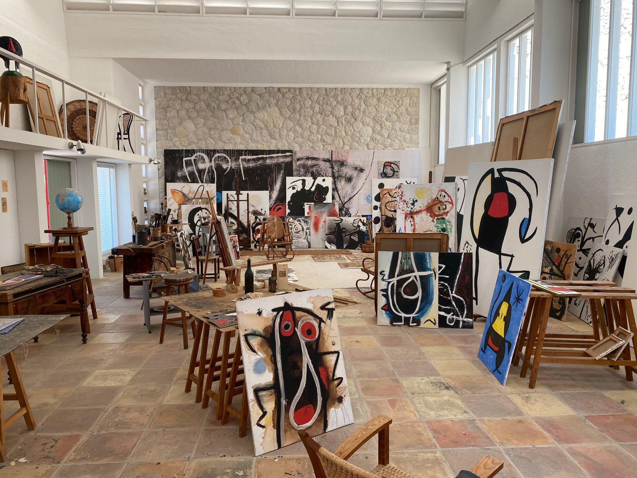 Joan Miró Foundation, Balearic Islands — Artist studio with terracotta floors, wooden chairs, textured stone wall, and abstract paintings in black, red, and blue tones.