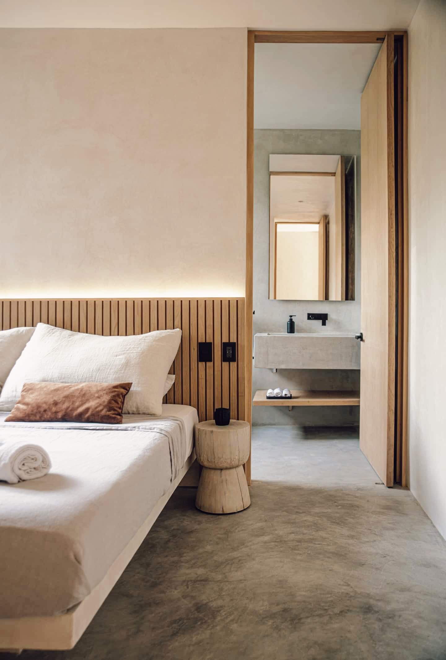 Villa Cava, Mexico — Minimalist bedroom suite with vertical wood-slat headboard, concrete sink vanity, and open ensuite visible through timber-framed doorway, bathed in warm ambient lighting.