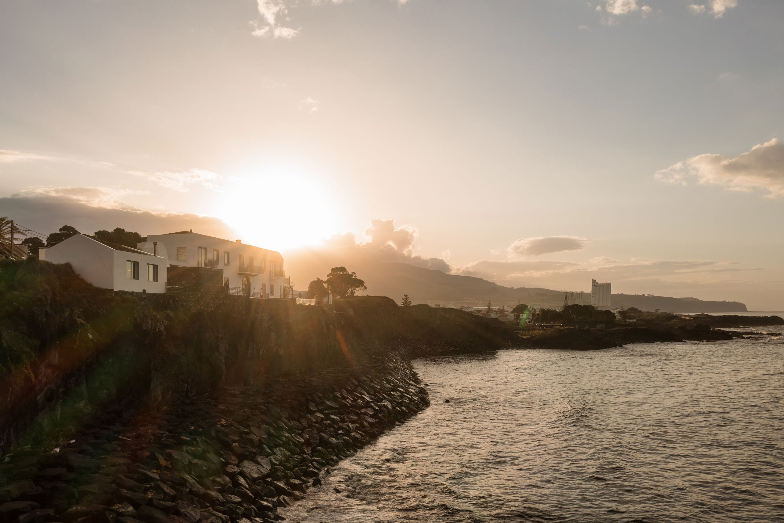 White Hotel, Europe — Clifftop modernist building at golden hour with rocky coastline, calm sea, and mountain backdrop under dramatic sunset sky.