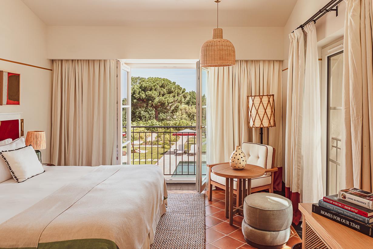 Hôtel Lou Pinet, Europe — Sunlit bedroom with terrace view, woven pendant light, linen curtains, wooden furnishings, and Mediterranean garden landscape.