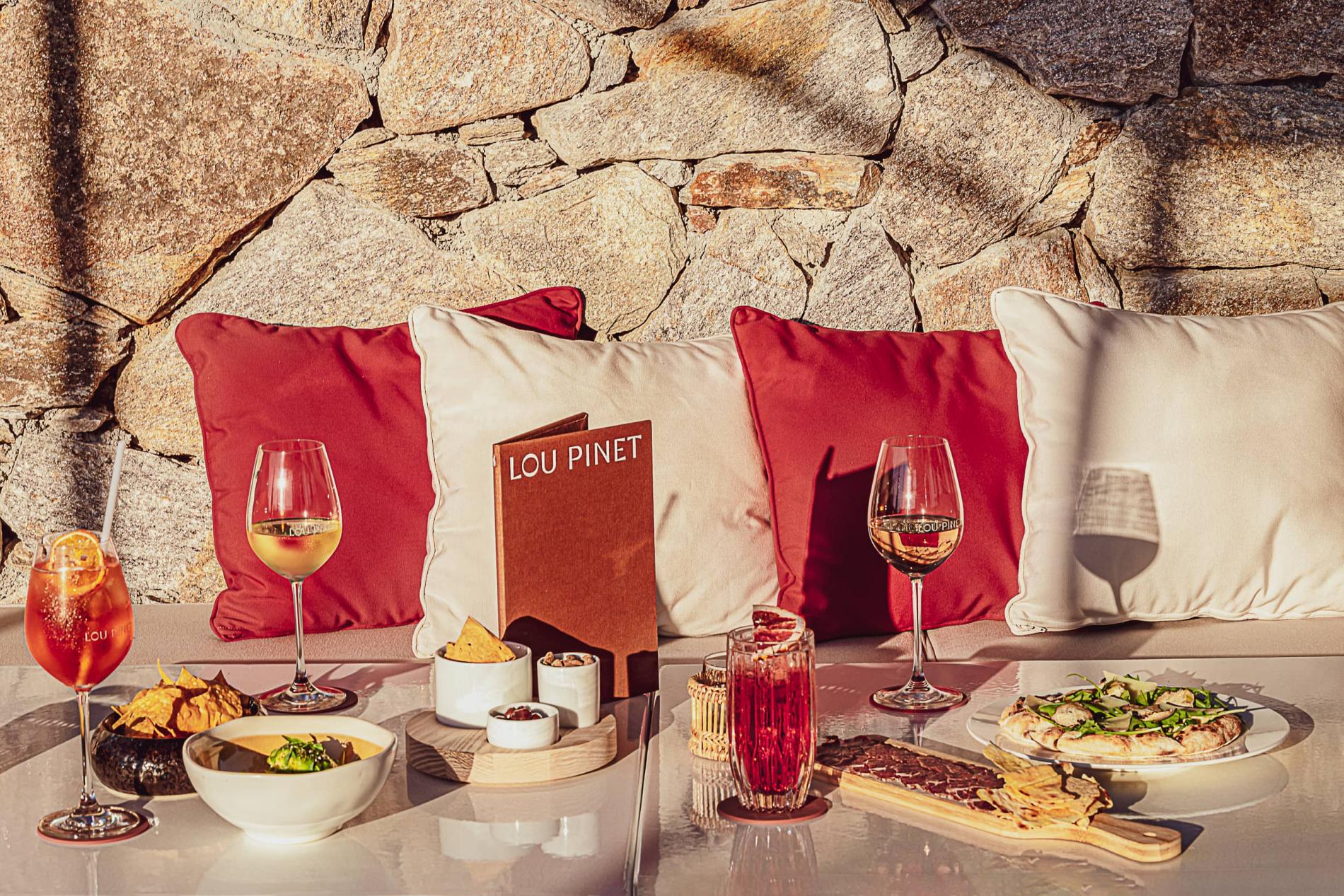 The Bar of Lou Pinet — Outdoor dining setup at Lou Pinet with red and cream cushions, wine glasses, charcuterie, and stone wall backdrop in Mediterranean sunlight.