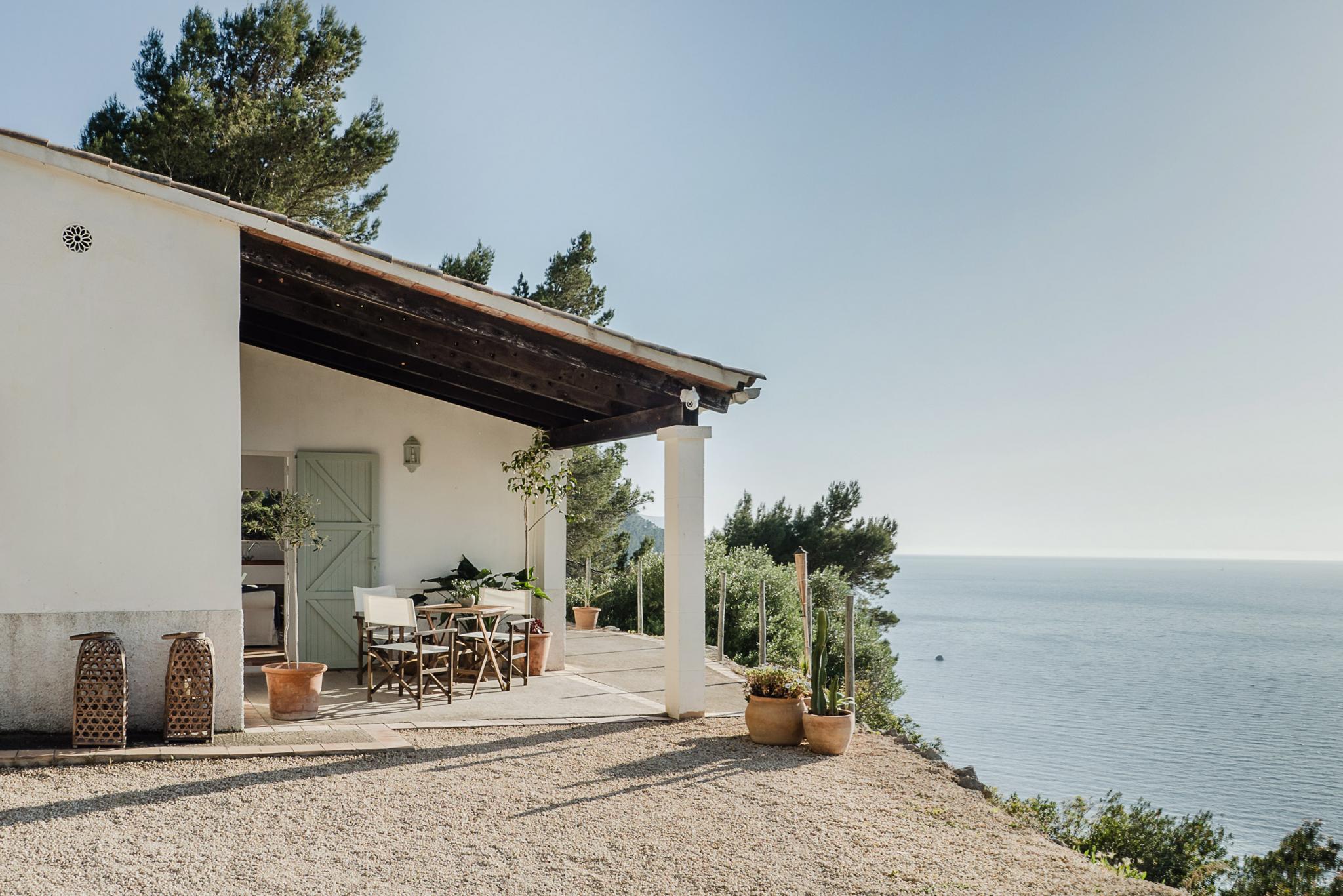 Hideaway, Balearic Islands, Spain — Modernist clifftop villa with dark timber pergola, whitewashed walls, and Mediterranean sea views through pine trees.
