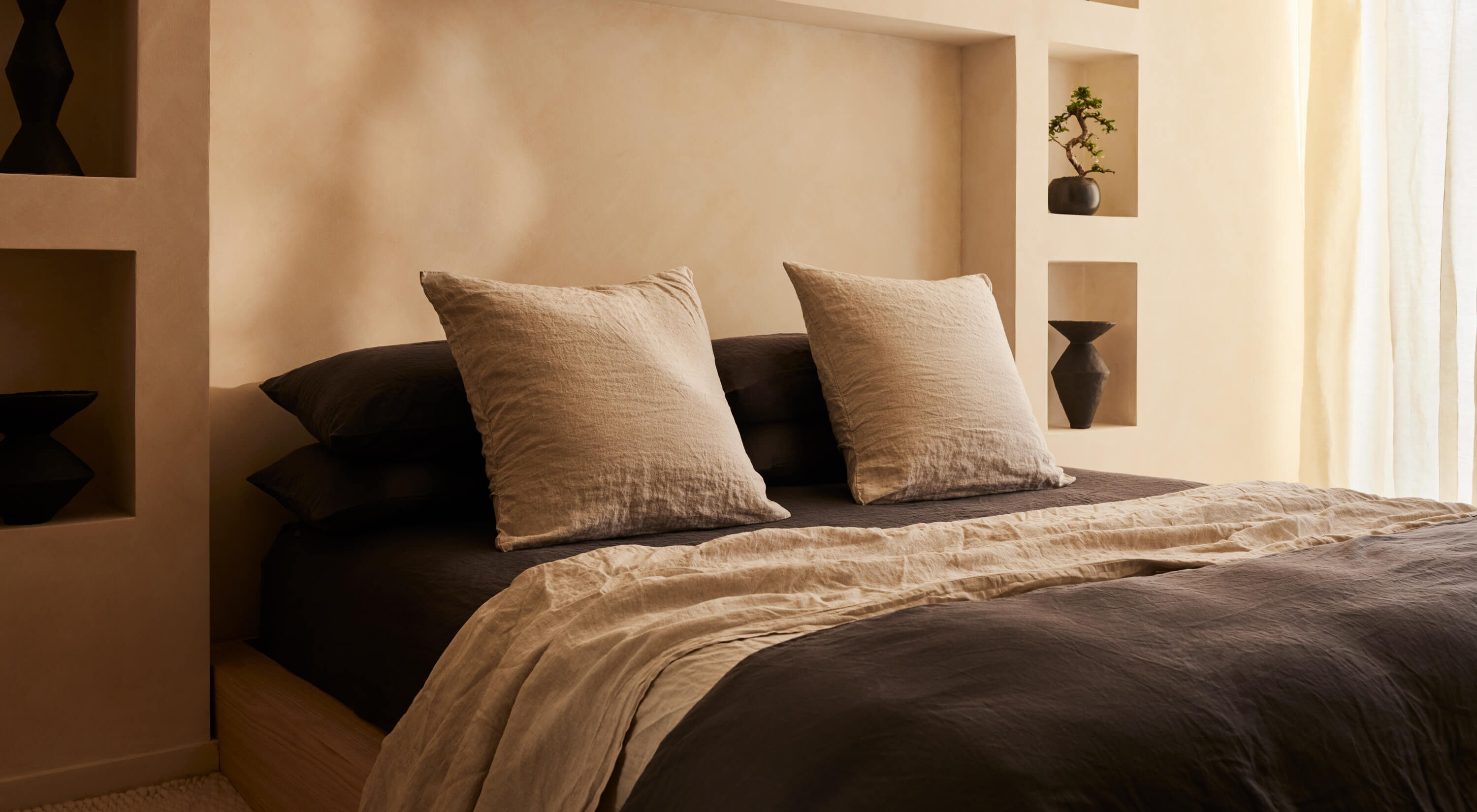 Arjé Home, North America — Minimalist bedroom with warm neutral walls, black bedding, linen pillows, natural light, and recessed shelving with ceramic vessels.
