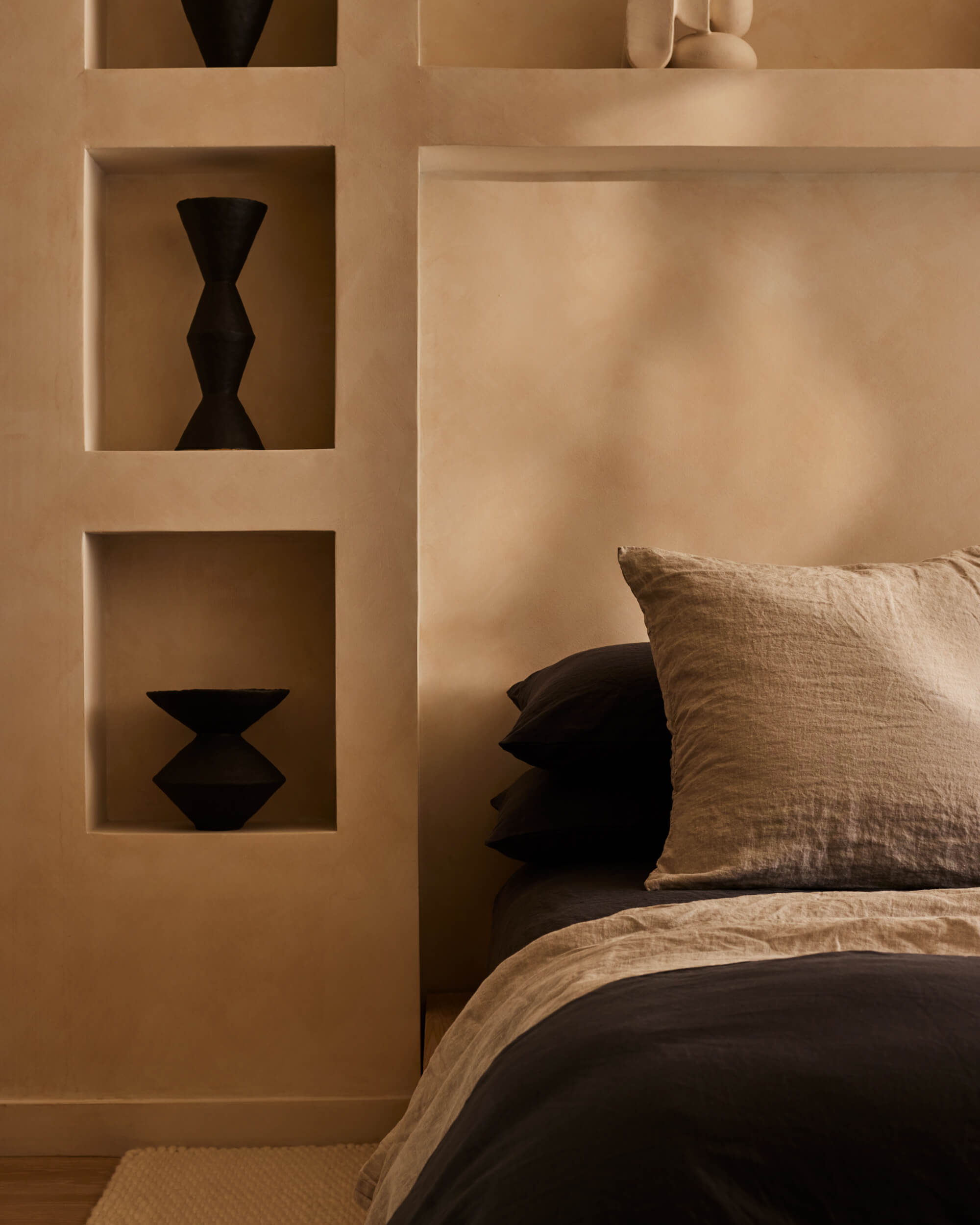 Arjé Home, North America — Minimalist bedroom with built-in wall niches displaying black sculptural vessels, neutral linen bedding, and warm ambient lighting creating serene modern aesthetic.