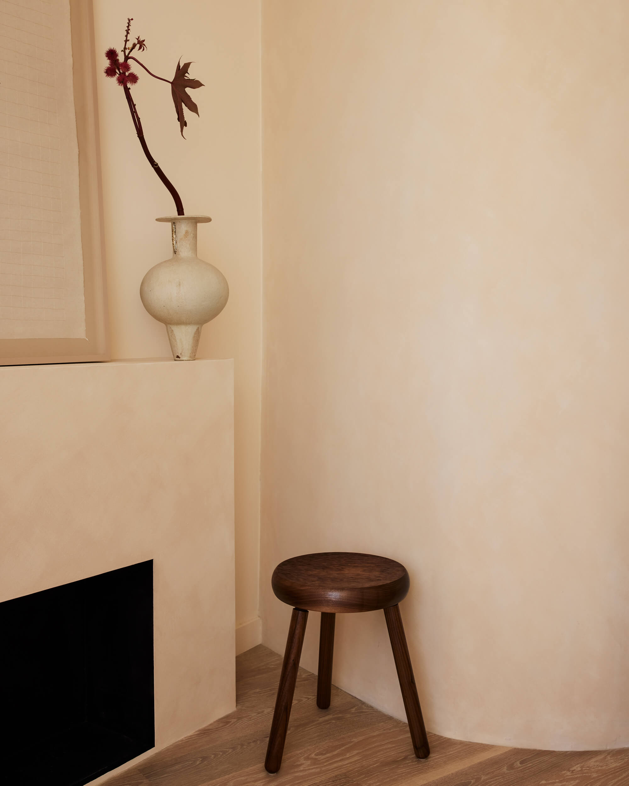 Arjé Home, North America — Minimalist interior with cream walls, wooden stool, pale ceramic vase holding dried burgundy botanicals, and black architectural element.