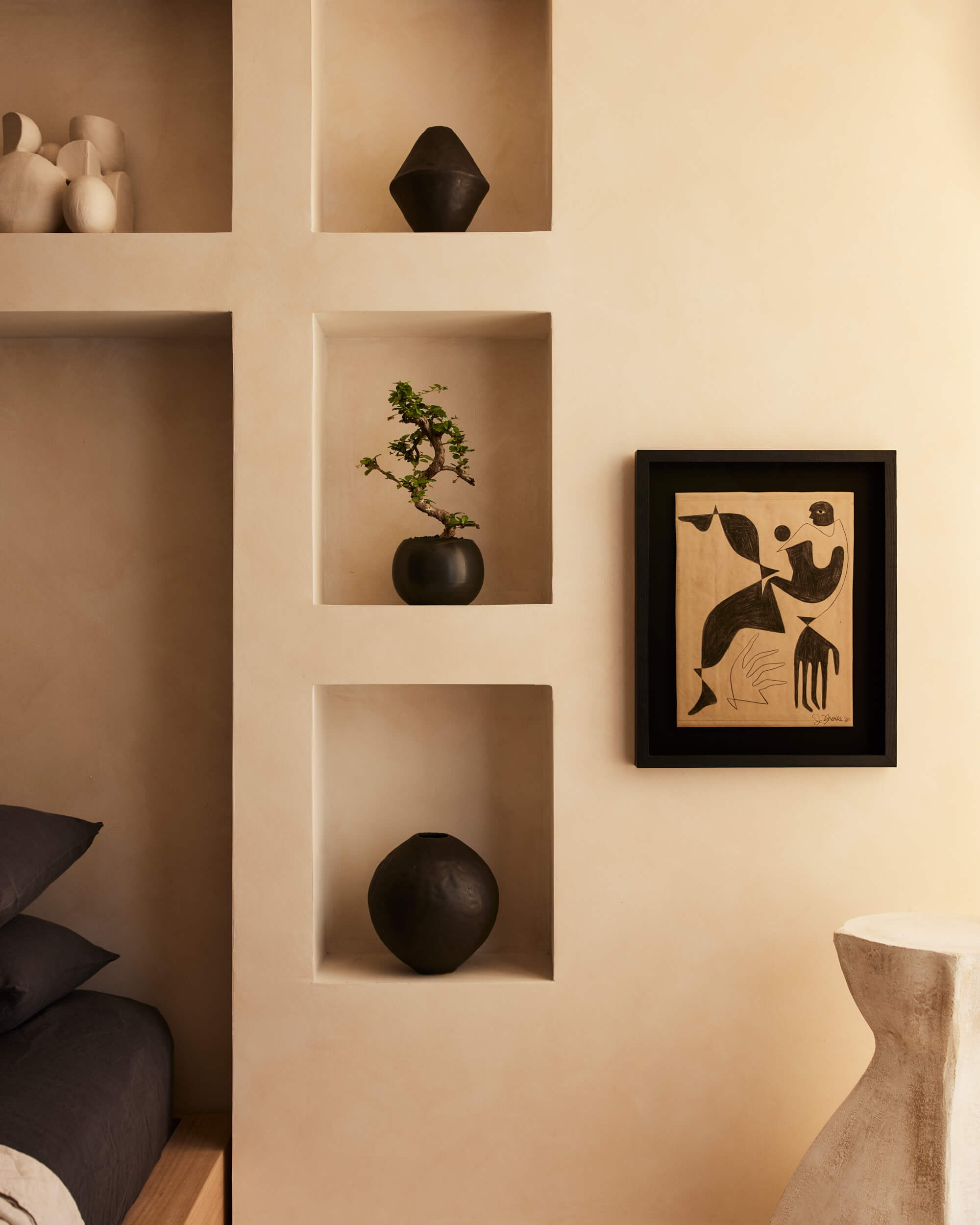 Arjé Home, North America — Minimalist wall niches display black ceramic vessels and bonsai against warm beige plaster, with framed abstract artwork and natural wood furniture.