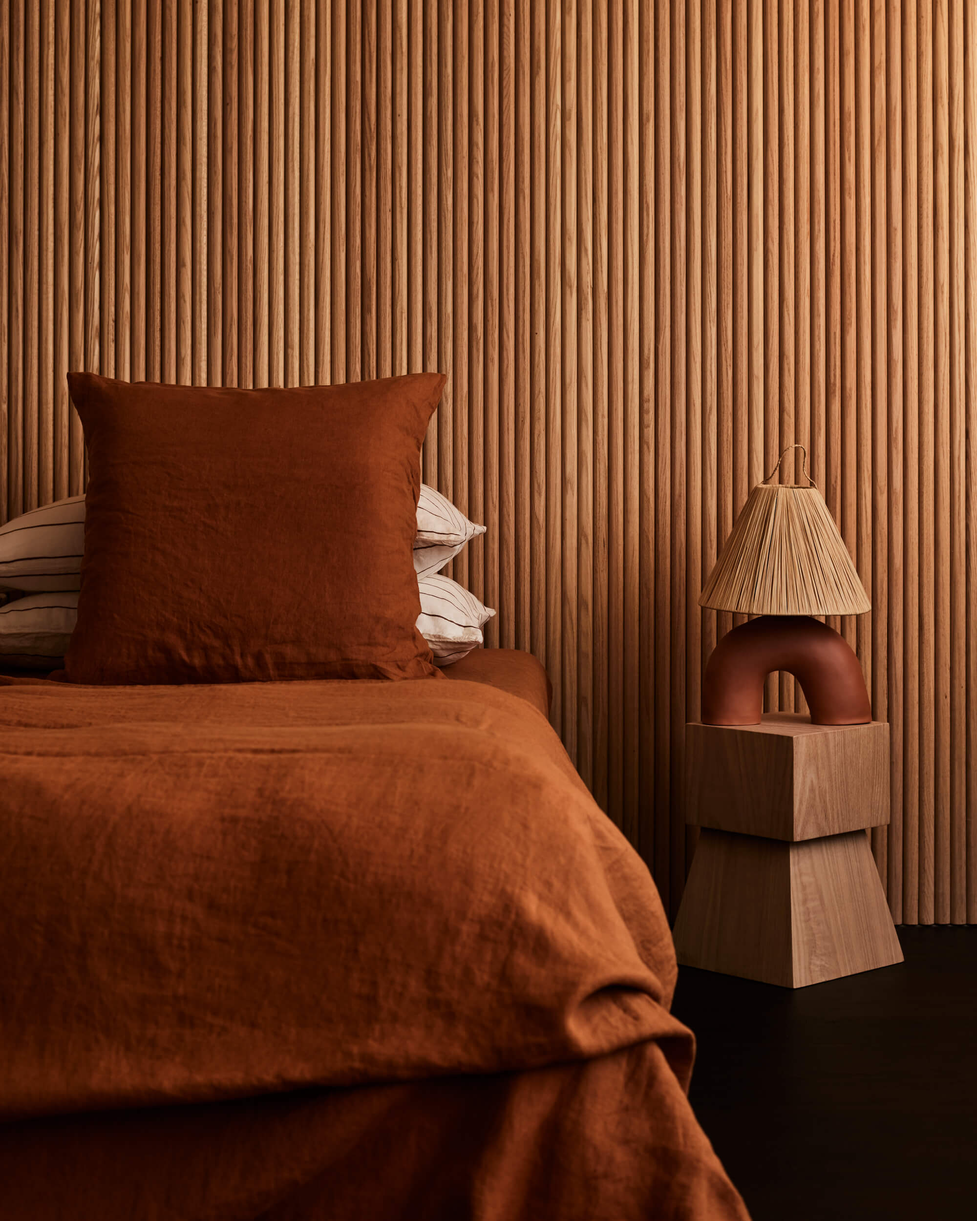 Arjé Home, North America — Terracotta linen bedding and rust-toned pillow against vertical wood-slat wall, ceramic sculptural nightstand with pleated shade, warm earthy minimalist bedroom.