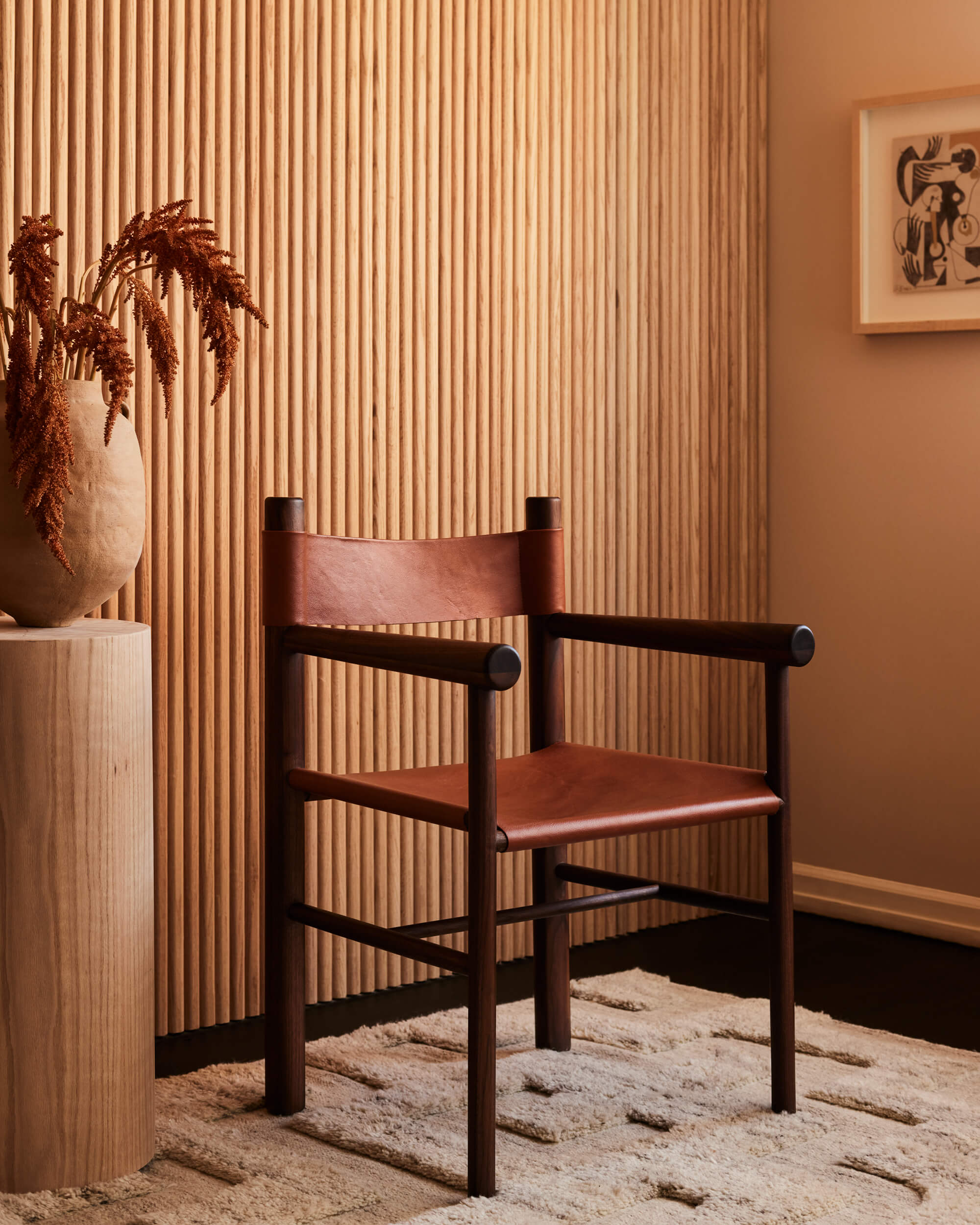 Arjé Home, North America — Dark wooden chair with cognac leather seat against fluted wall paneling, styled with terracotta vessel and minimalist decor.